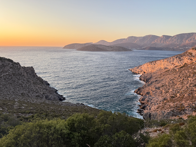 Sunset over a coastal landscape with hills and rocky cliffs surrounding a calm ocean.