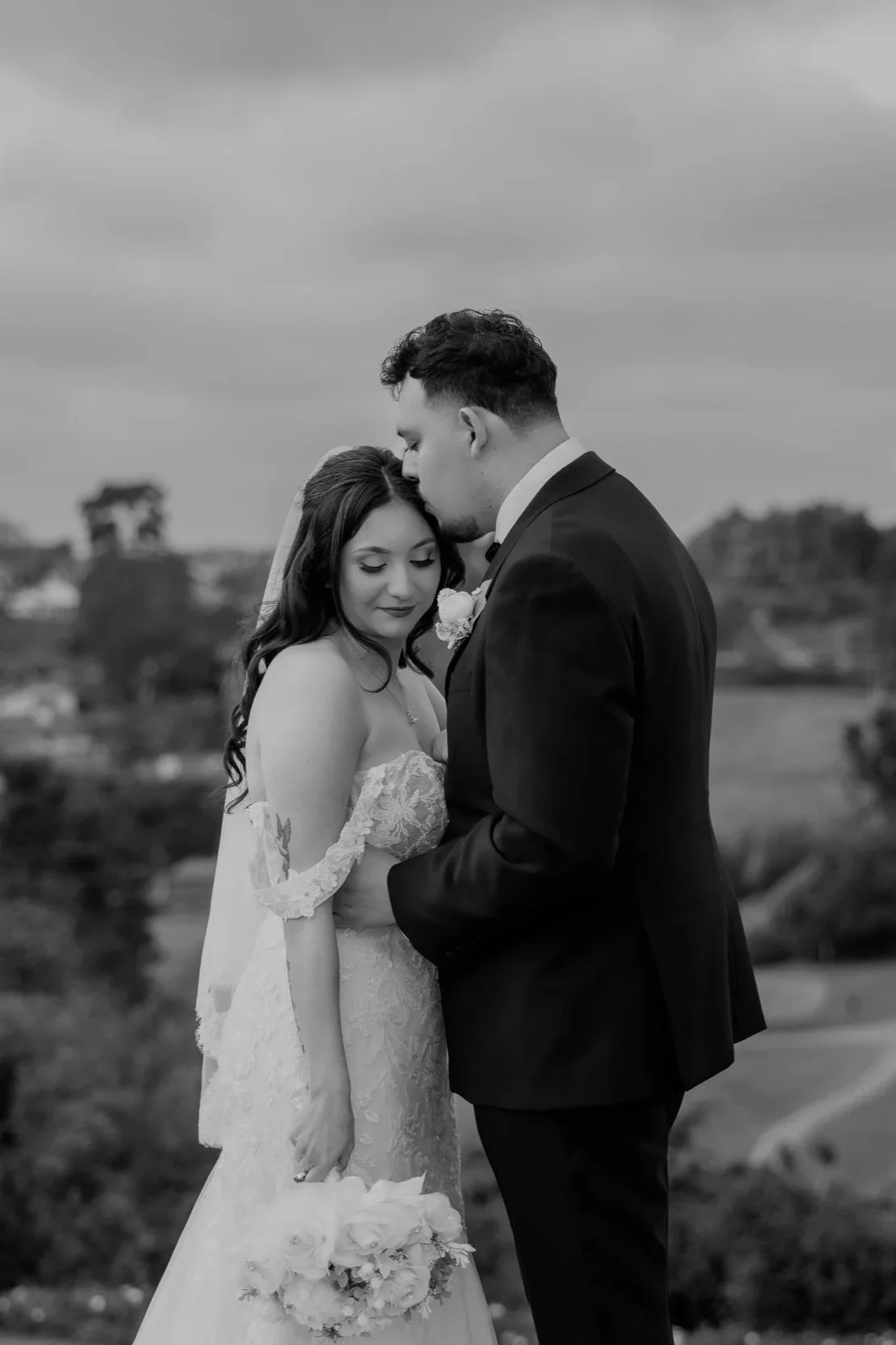 A black and white photograph of a bride and groom on their wedding day. The groom is kissing the bride's forehead while she holds a bouquet of flowers.