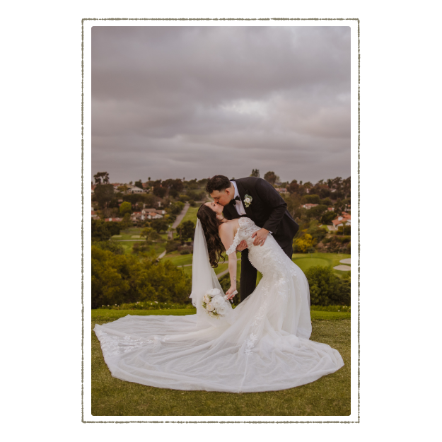 Bride in a white wedding dress and veil with a bouquet, and groom in a black tuxedo, sharing a kiss outdoors with a scenic green landscape and cloudy sky in the background.
