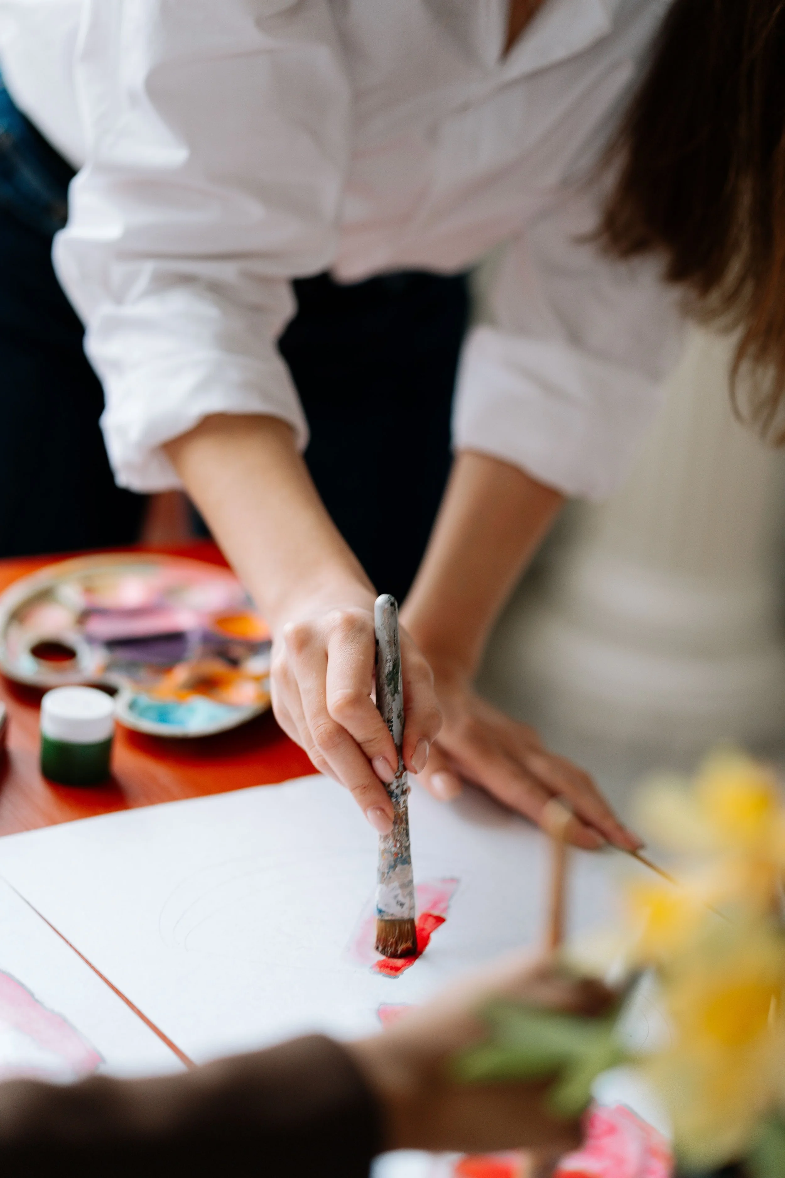 A person wearing a white shirt painting on paper with a brush, with artist supplies including paint bottles and a palette nearby.