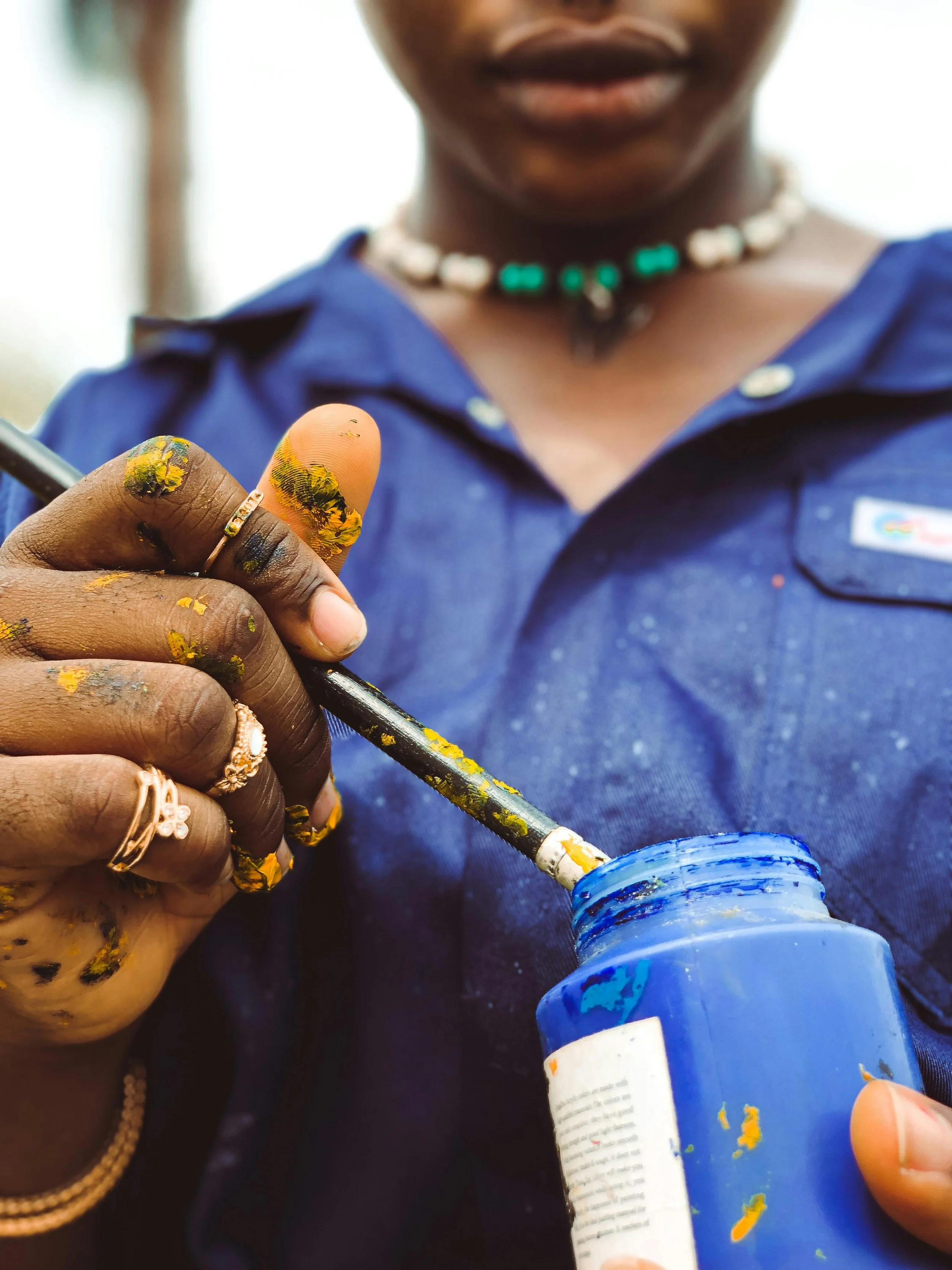 A person with dark skin, wearing a blue shirt and jewelry, is holding and dipping a paintbrush into a blue paint jar. Their hands and fingers are covered with yellow paint.