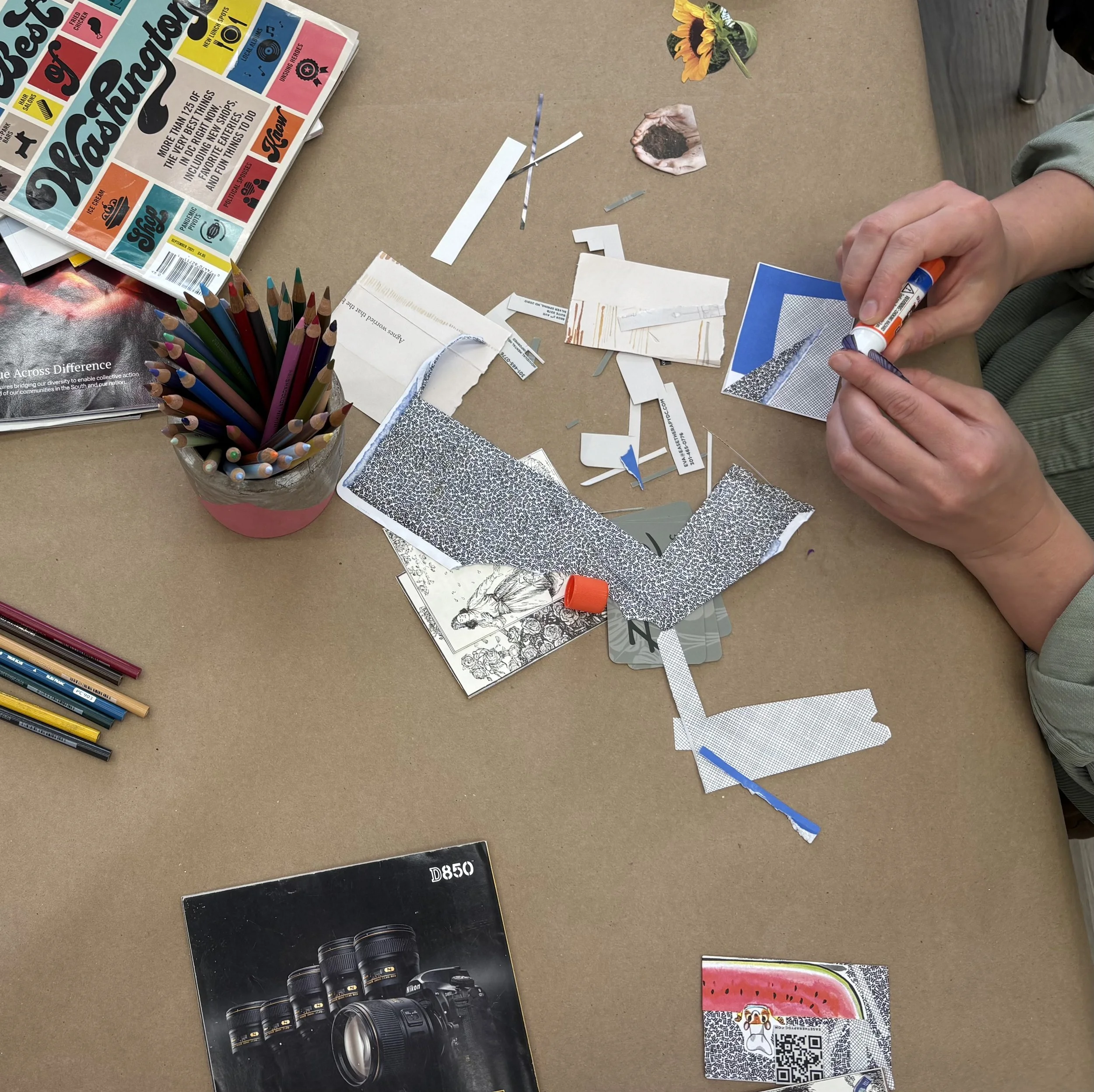 A table covered with scraps of paper, a container of colorful colored pencils, a camera brochure, and a person’s hand holding a glue stick. Part of a magazine and some booklets are also visible.