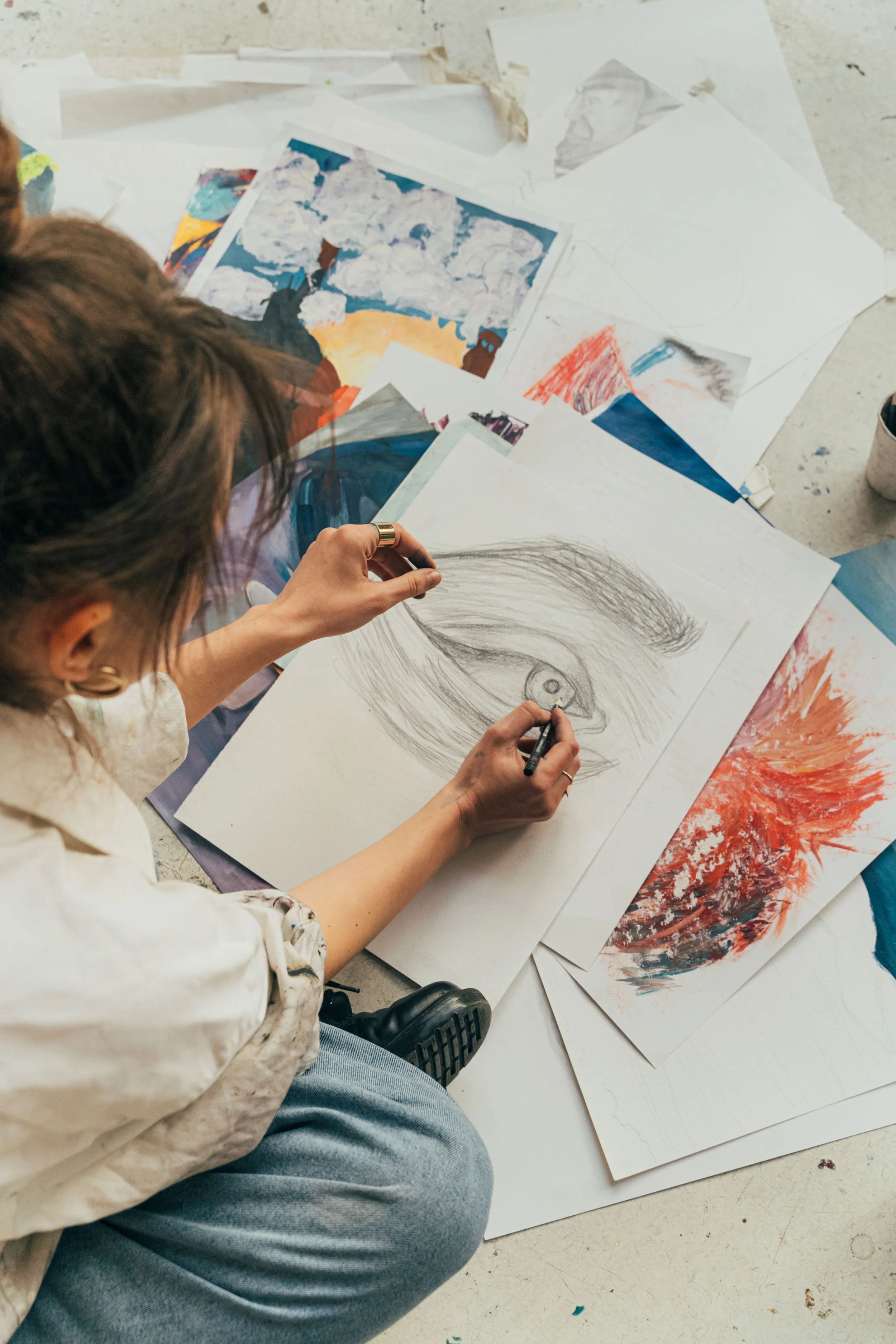Person sketching a detailed eye on paper with several other artworks and sketches scattered on the floor around.