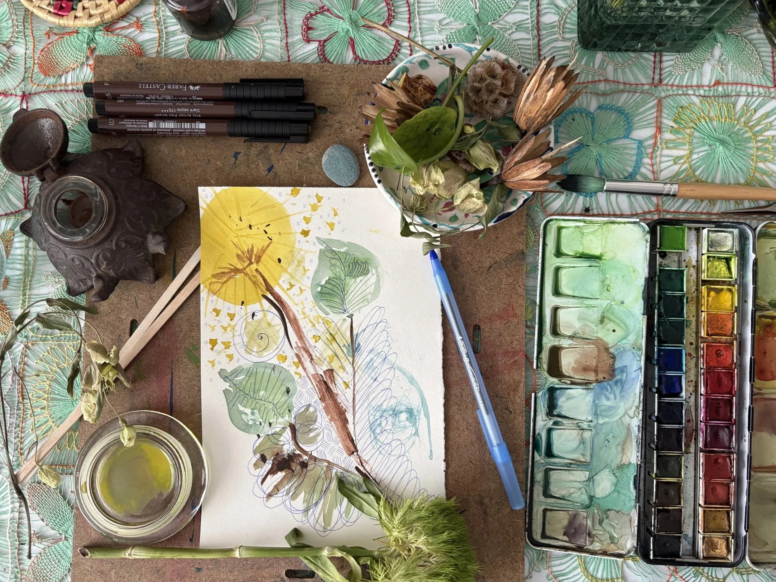 A watercolor painting in progress on paper, showcasing a sun, branches, and leaves, surrounded by art supplies including watercolor paints, brushes, markers, and dried plants on a textured surface.