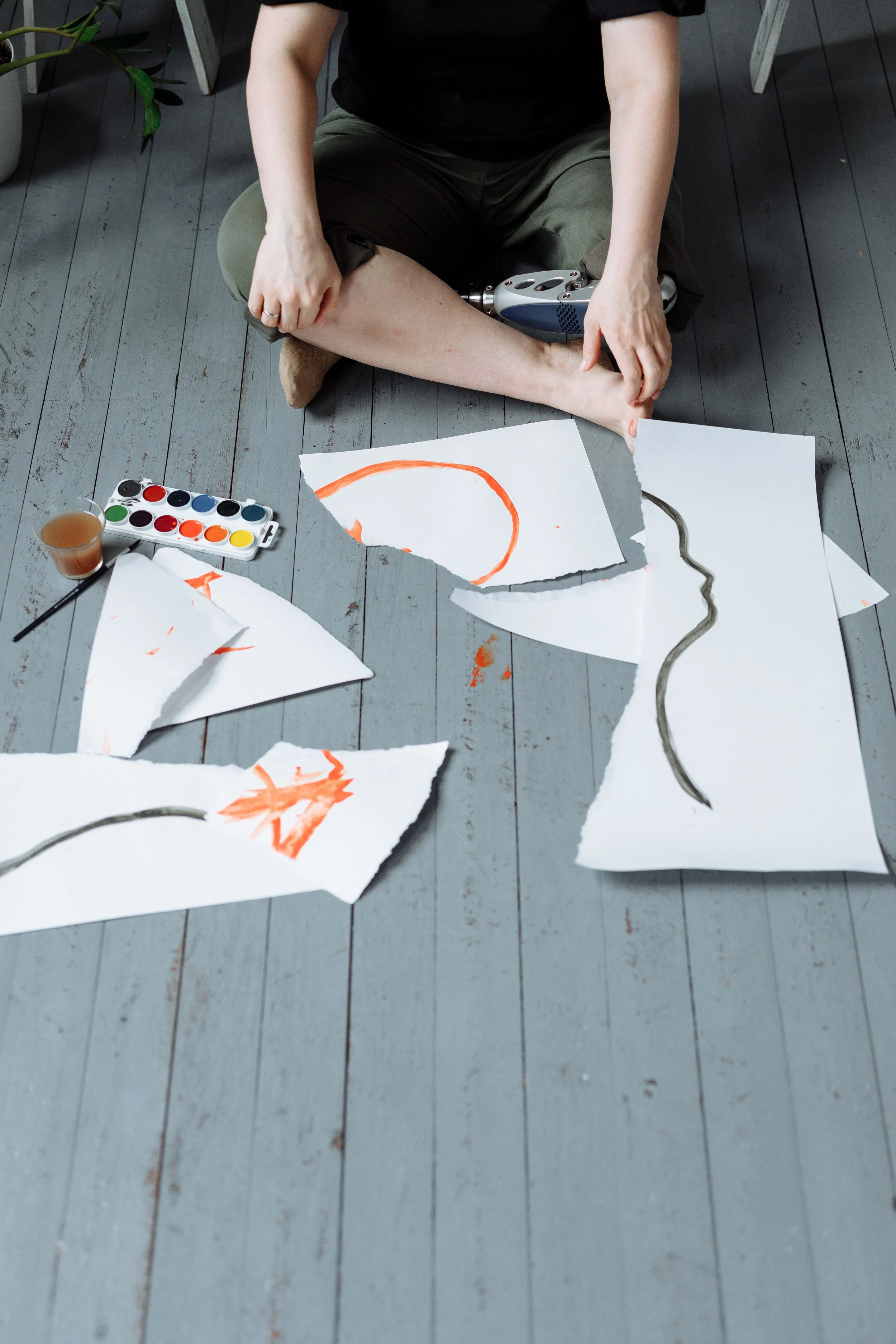 Person sitting on gray wooden floor, holding a prosthetic leg, surrounded by torn white papers with orange and black markings, watercolor paints, a glass of tea, and a paintbrush.