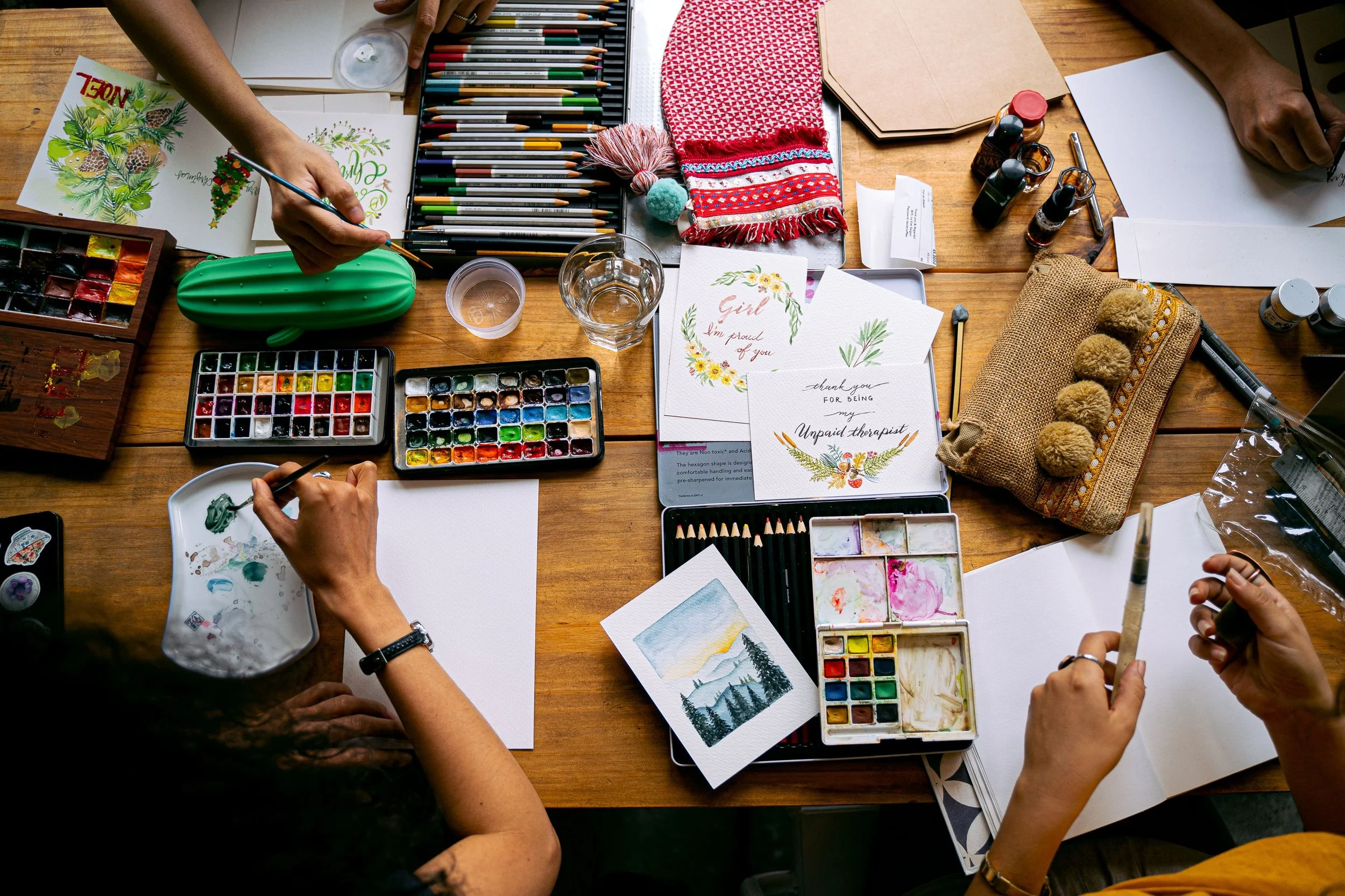 A table with art supplies including watercolors, colored pencils, and brushes. Several people are painting and drawing, with finished cards and artwork. Decorative items include a cactus-shaped pencil case, a red scarf, and greeting cards with floral designs and messages.