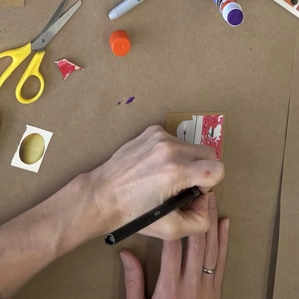 A person is using a black marker to write on a small card or photo that has red and white paint splatters. The table has various art supplies including scissors with yellow handles, glue or paint bottles, and small torn pieces of paper, with a hand and part of the person's finger with a wedding ring visible.