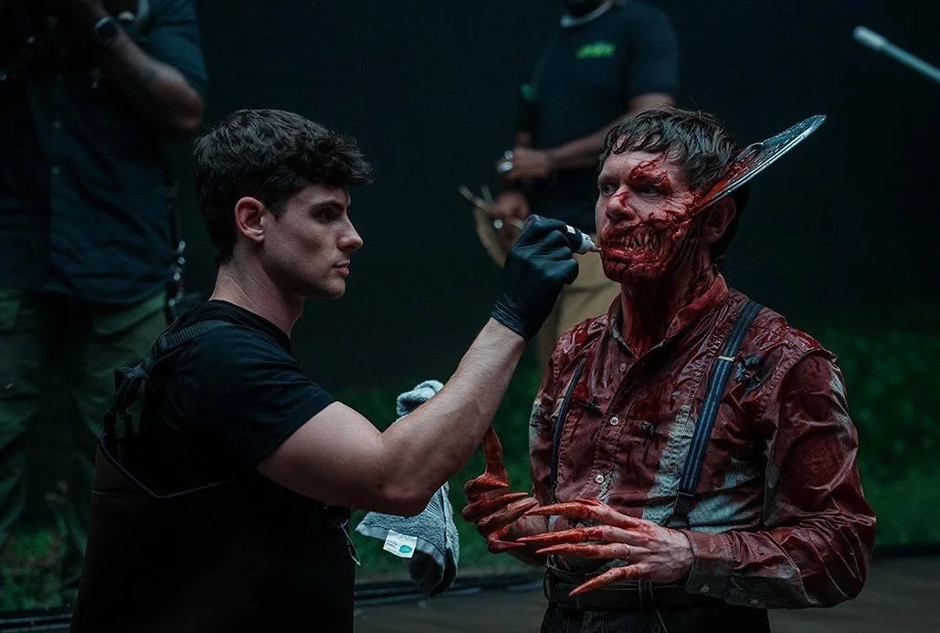 Behind the scenes of a movie or TV show with a makeup artist applying special effects to an actor's face to look like a zombie.