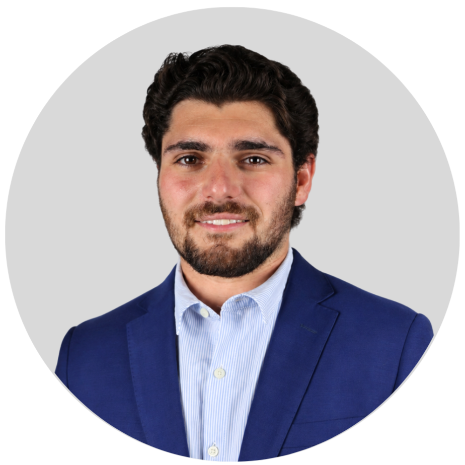 Headshot of a man with dark brown curly hair and a beard, wearing a blue blazer and light blue collared shirt, smiling against a plain gray background.