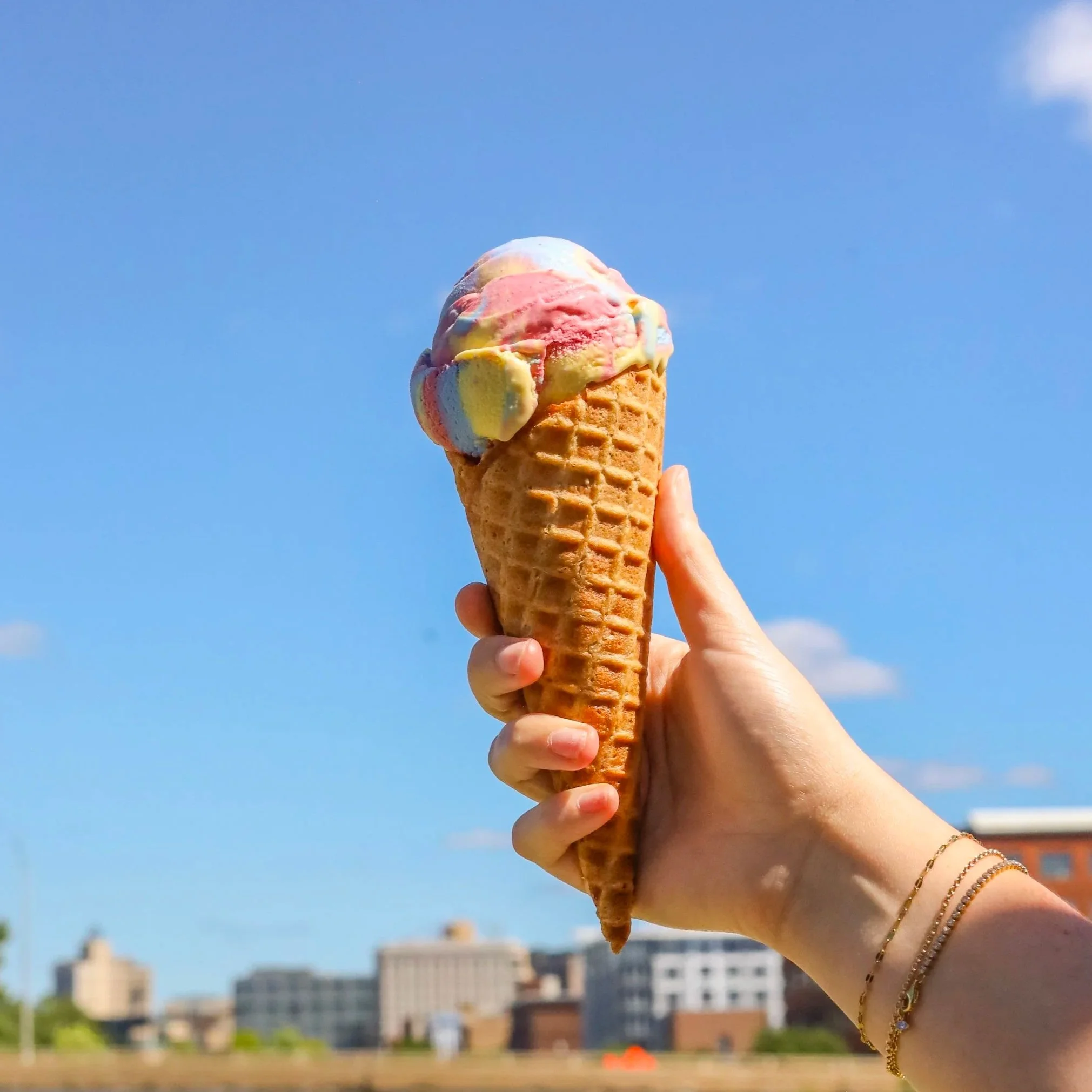 A colorful scoop of ice cream served in a waffle cone being held high with a blue sky in the background.