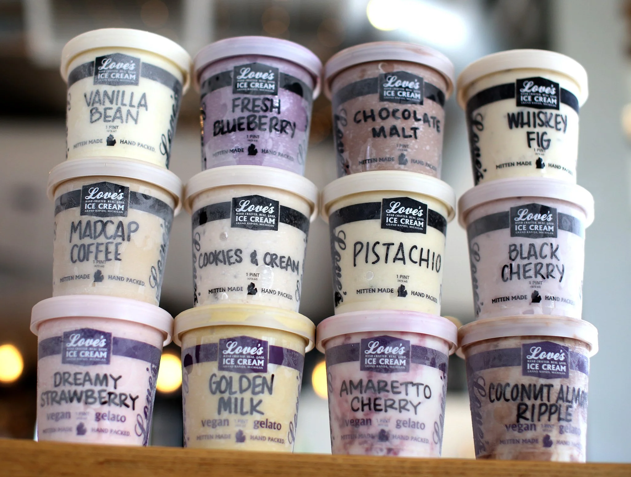 Stack of fifteen ice cream pints with various flavors labeled, including vanilla bean, fresh blueberry, chocolate malt, whiskey fig, madcap coffee, cookies & cream, pistachio, black cherry, dreamy strawberry, golden milk, amaretto cherry, and coconut
