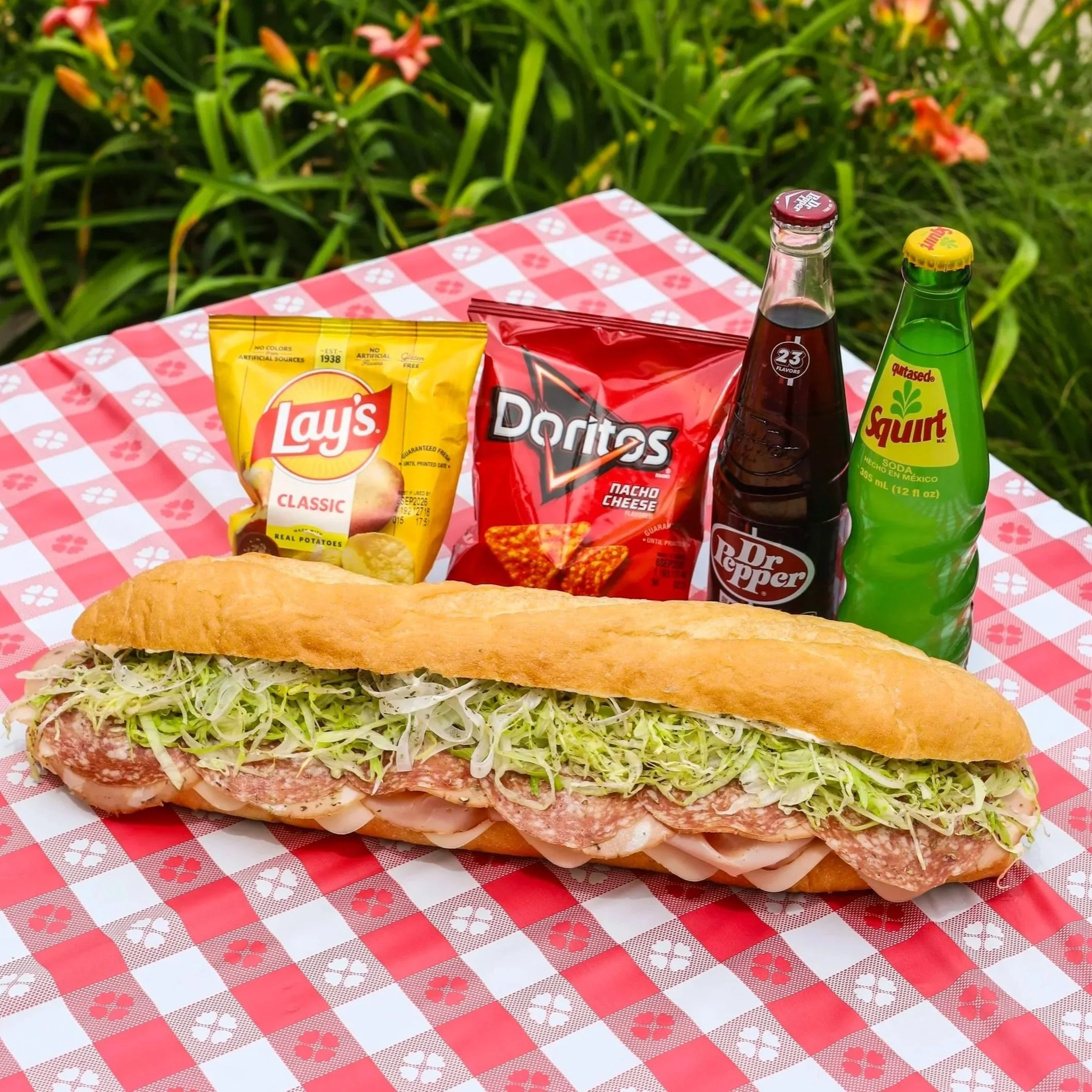 A huge sub sandwich sitting on a tablecloth outside next to two craft sodas and two bags of chips.