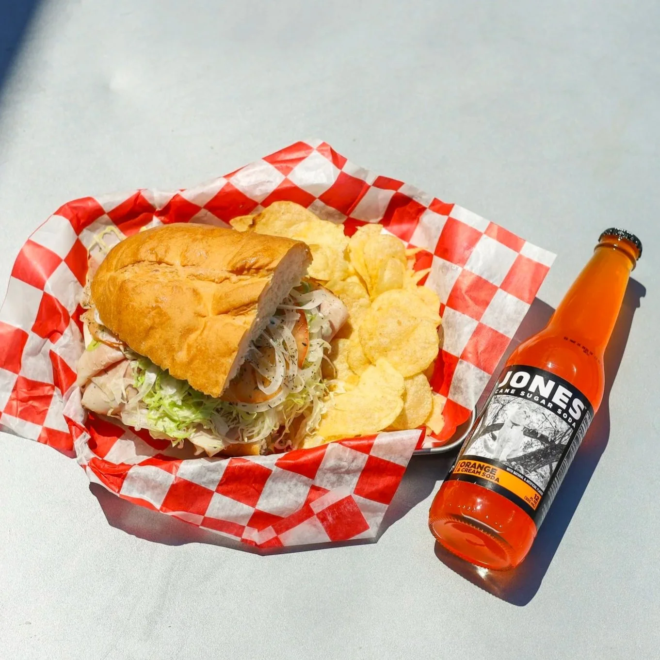 A plate with a sandwich containing lettuce, tomato, and turkey, served with potato chips, next to a bottle of Goody Berry Cream soda.