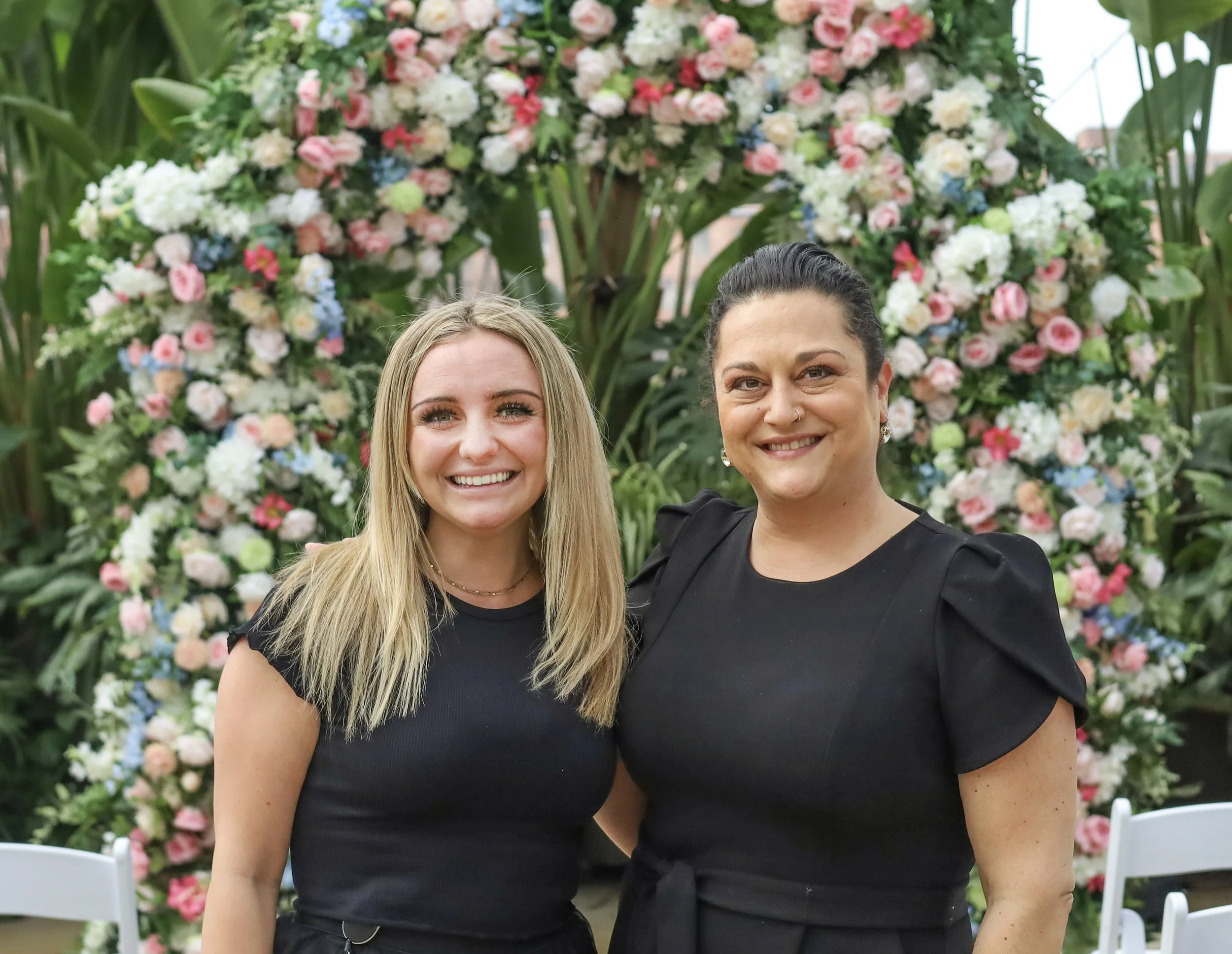 Two women smiling at a floral event with a backdrop of colorful flowers arranged in an arch.