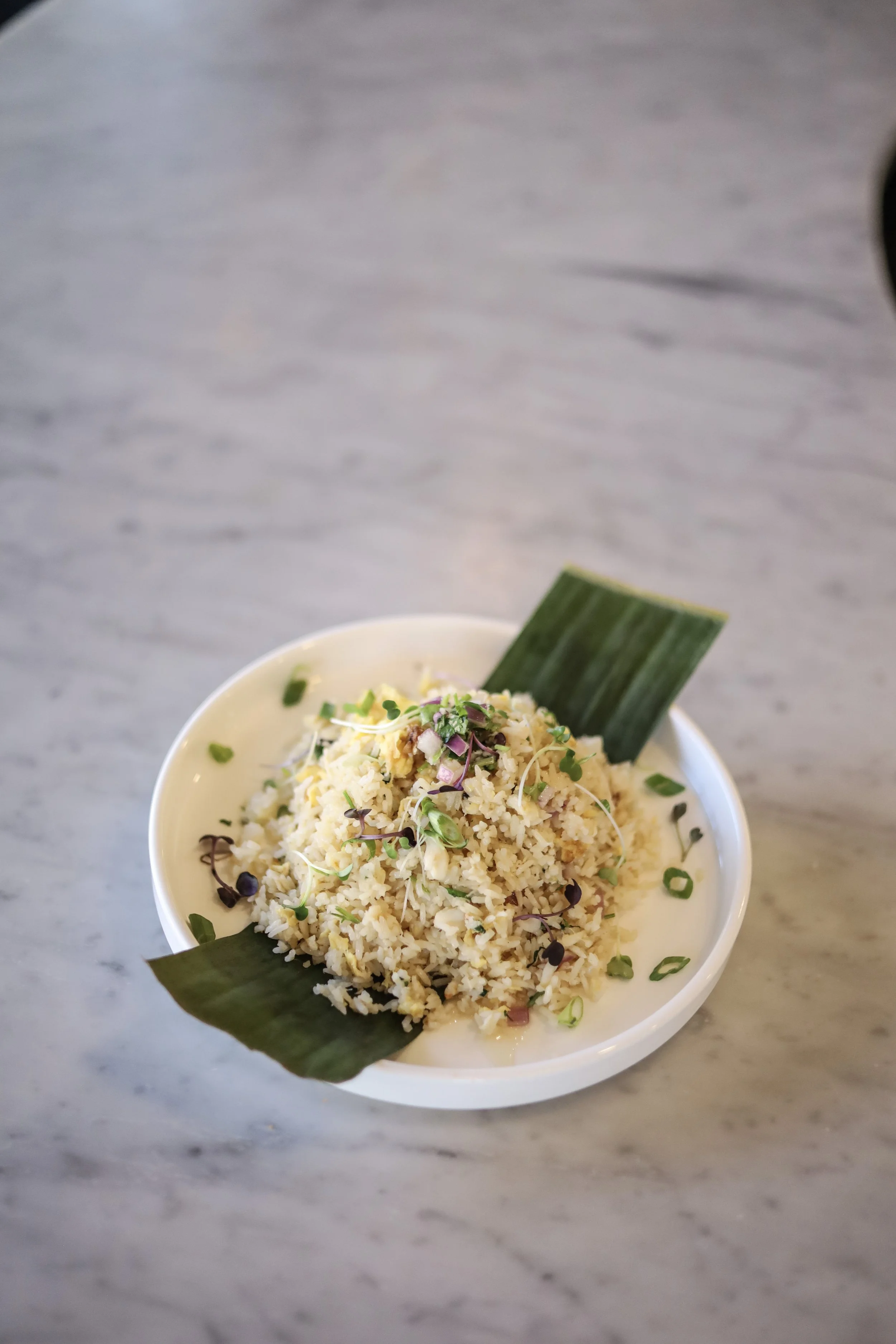 A bowl of crab fried rice garnished with microgreens and herbs, with two large green leaves inside the bowl, placed on a grey marble surface.