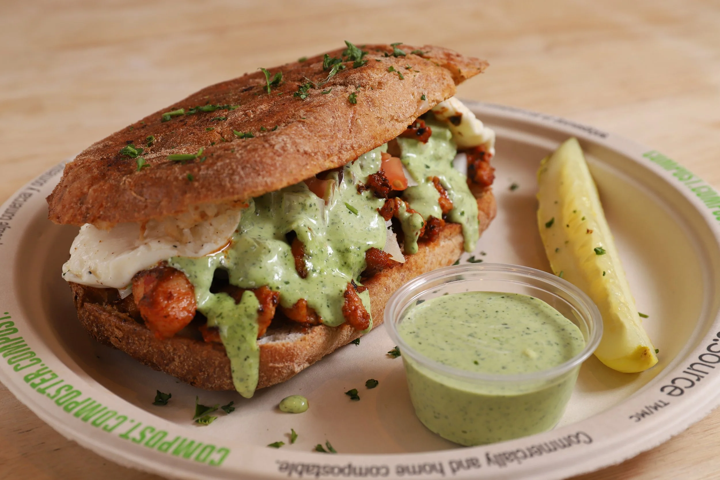Gourmet breakfast sandwich with spicy chicken and avocado sauce in a whole-grain bun, served with pickle and green sauce on side.