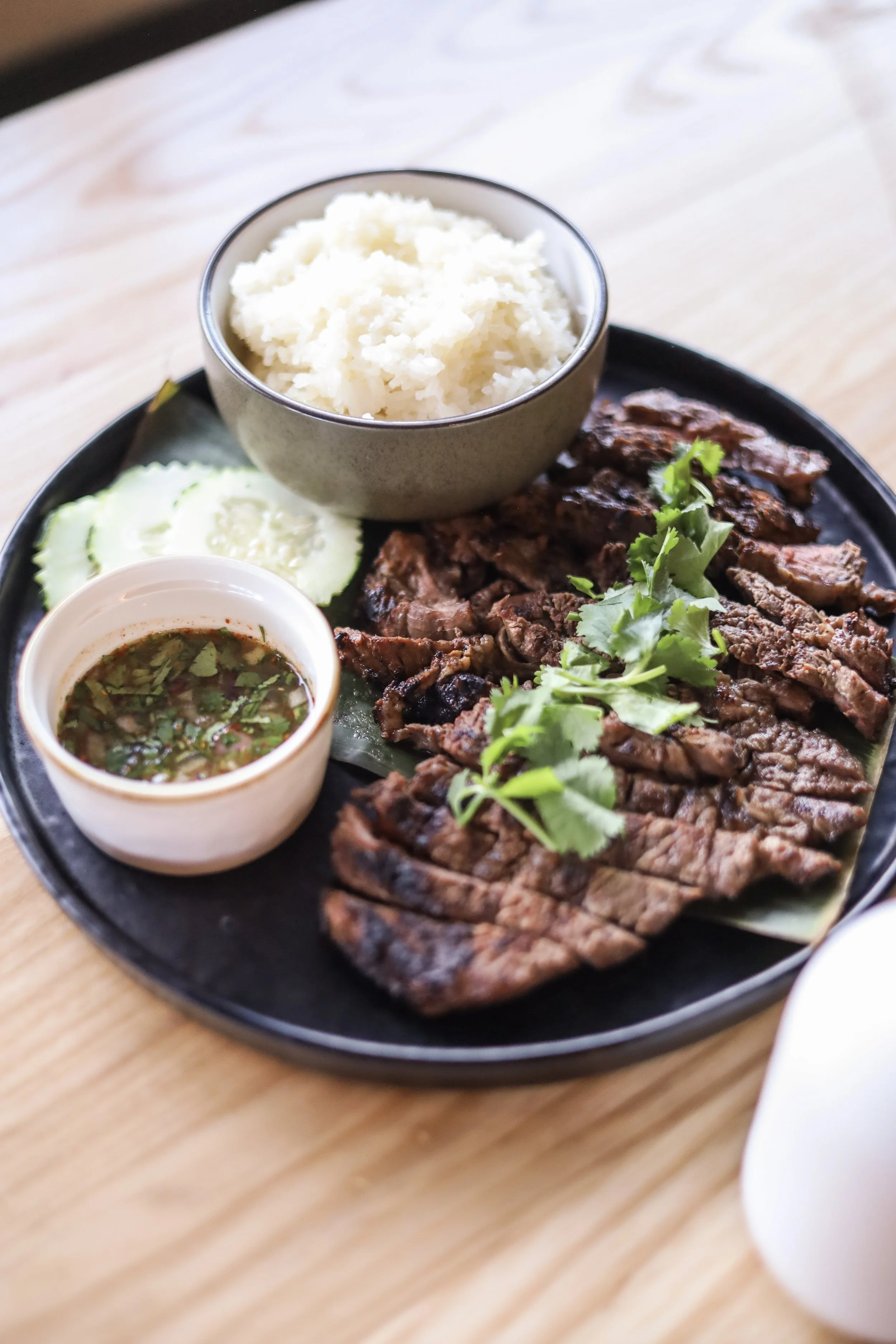 Plate of grilled meat with sliced steak, white rice in a bowl, cucumber slices, and a small bowl of dipping sauce, all garnished with fresh cilantro on a black tray.