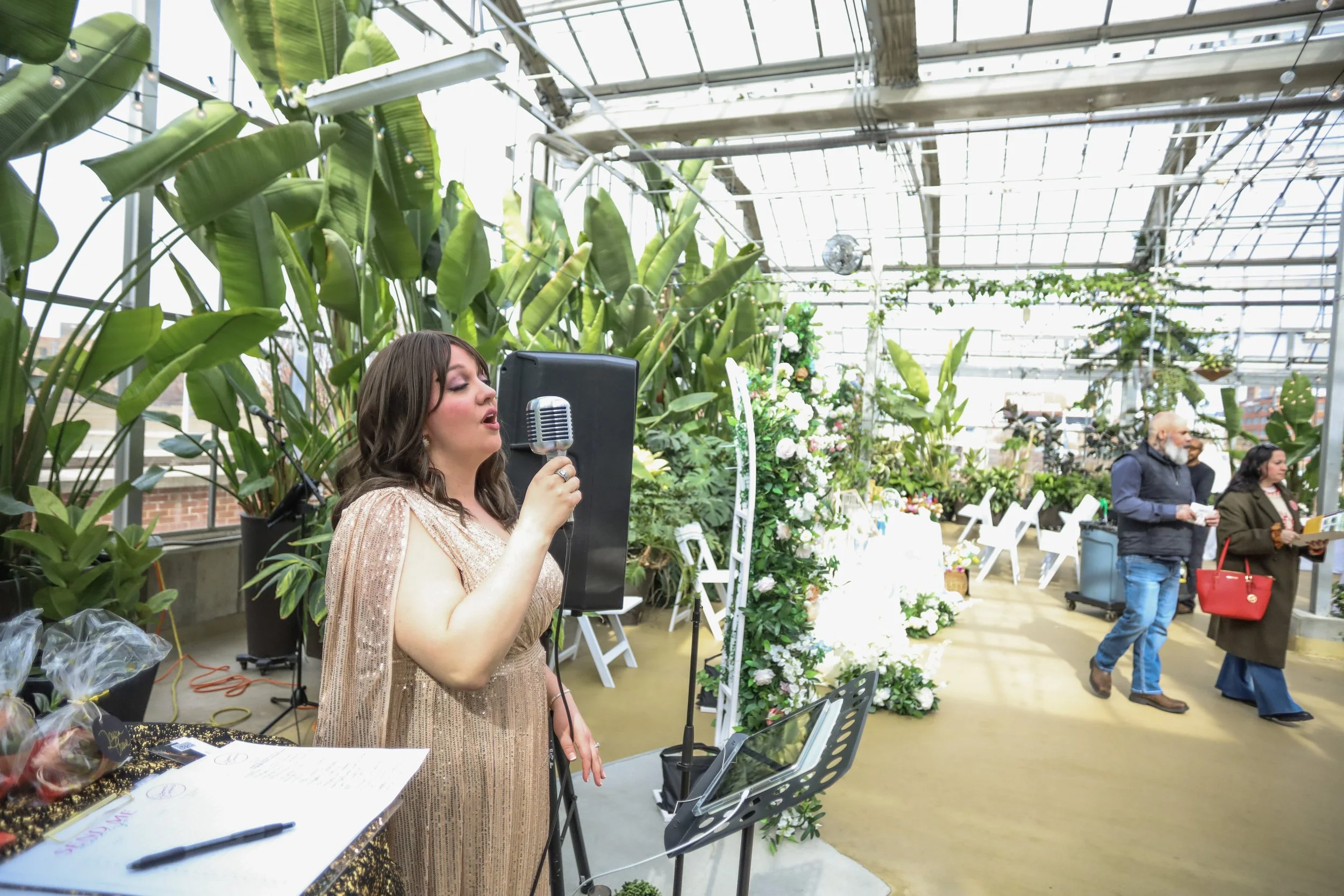 A woman singing into a microphone at a floral event inside a greenhouse with green plants and flowers, and a few people in the background.