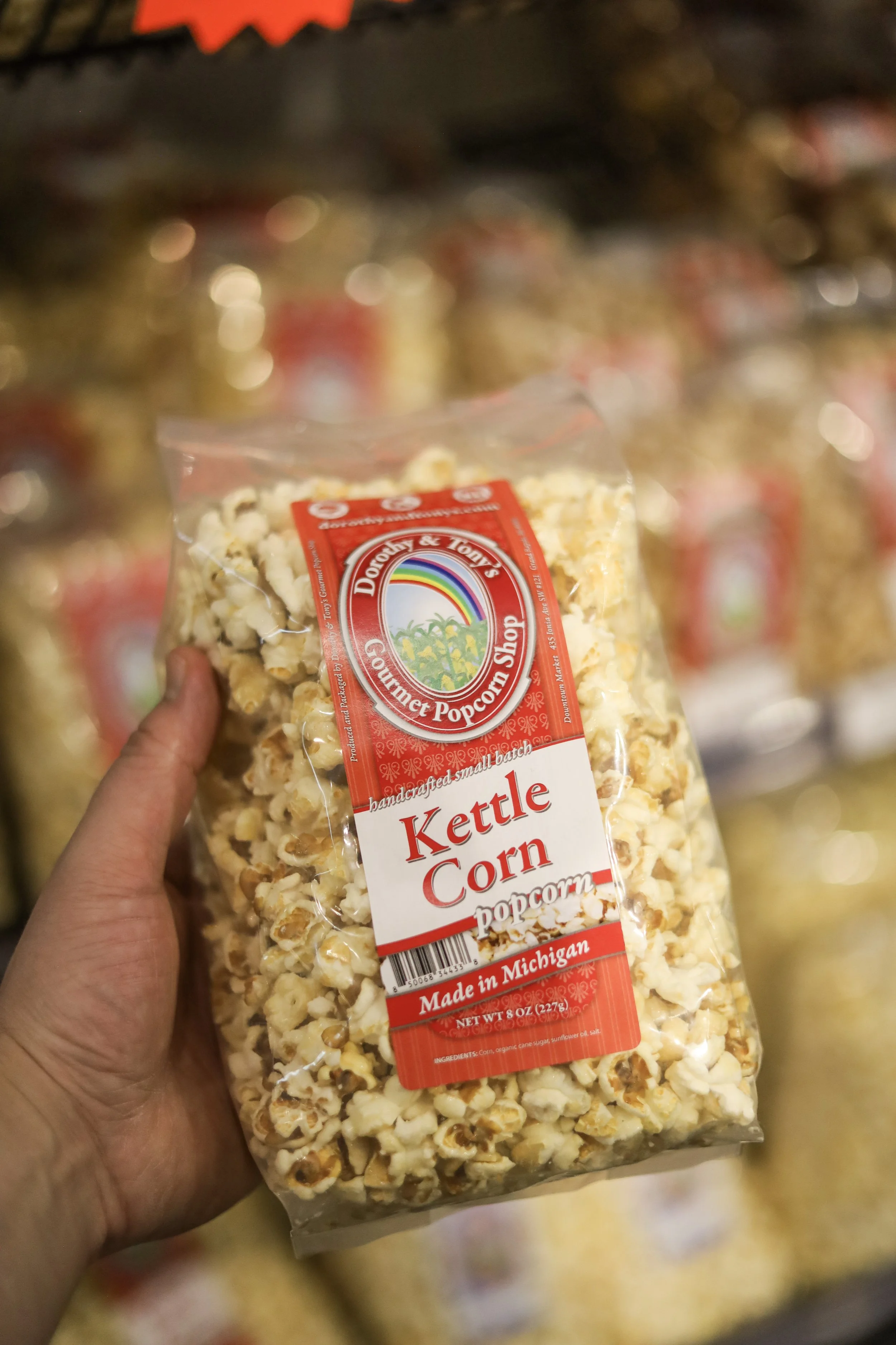 Hand holding a bag of kettle corn popcorn in a store aisle. The bag has a red and white label with a rainbow and palm trees, indicating it is from Dorothy & Tony's Gourmet Popcorn Shop, made in Michigan.