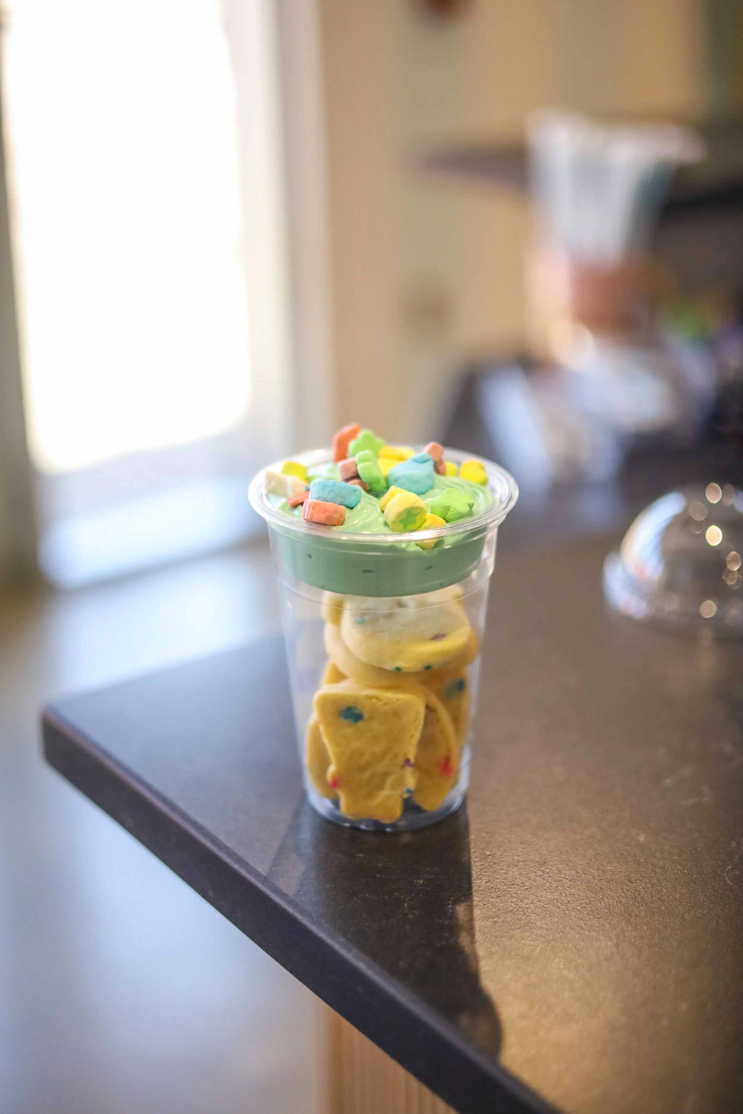 A clear plastic cup filled with colorful cookie pieces, green frosting, and sprinkles on top, placed on a dark countertop.