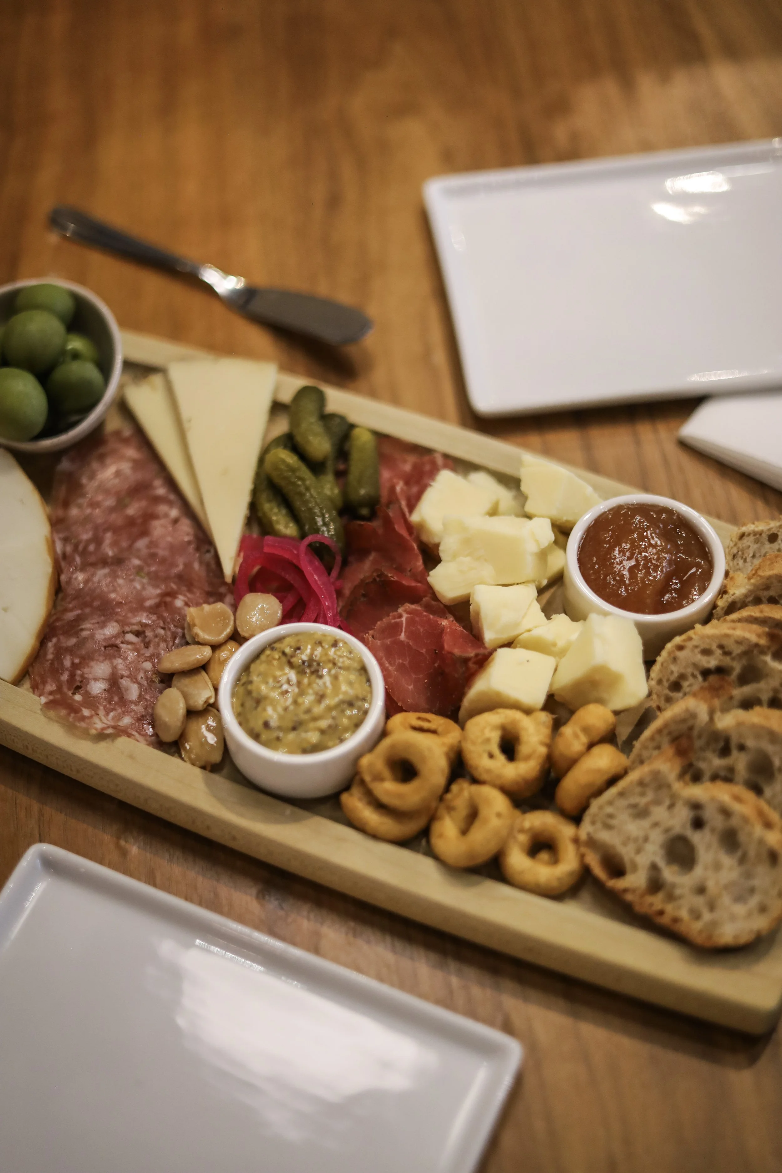A charcuterie board with various cheeses, meats, pickles, nuts, and bread, served with small bowls of mustard and jam, on a wooden table.
