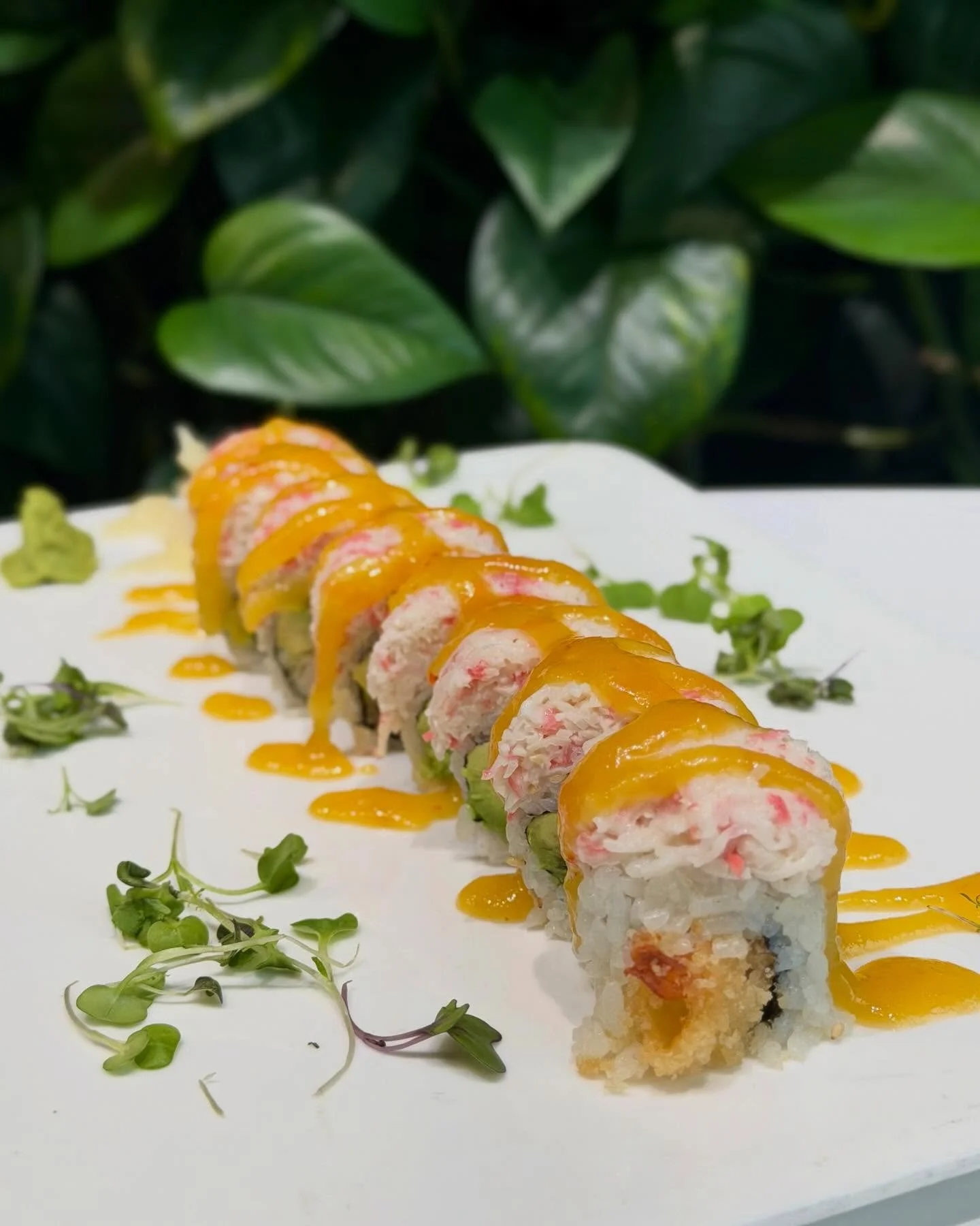 Sliced sushi roll drizzled with orange sauce, garnished with microgreens on a white plate with leafy green background.