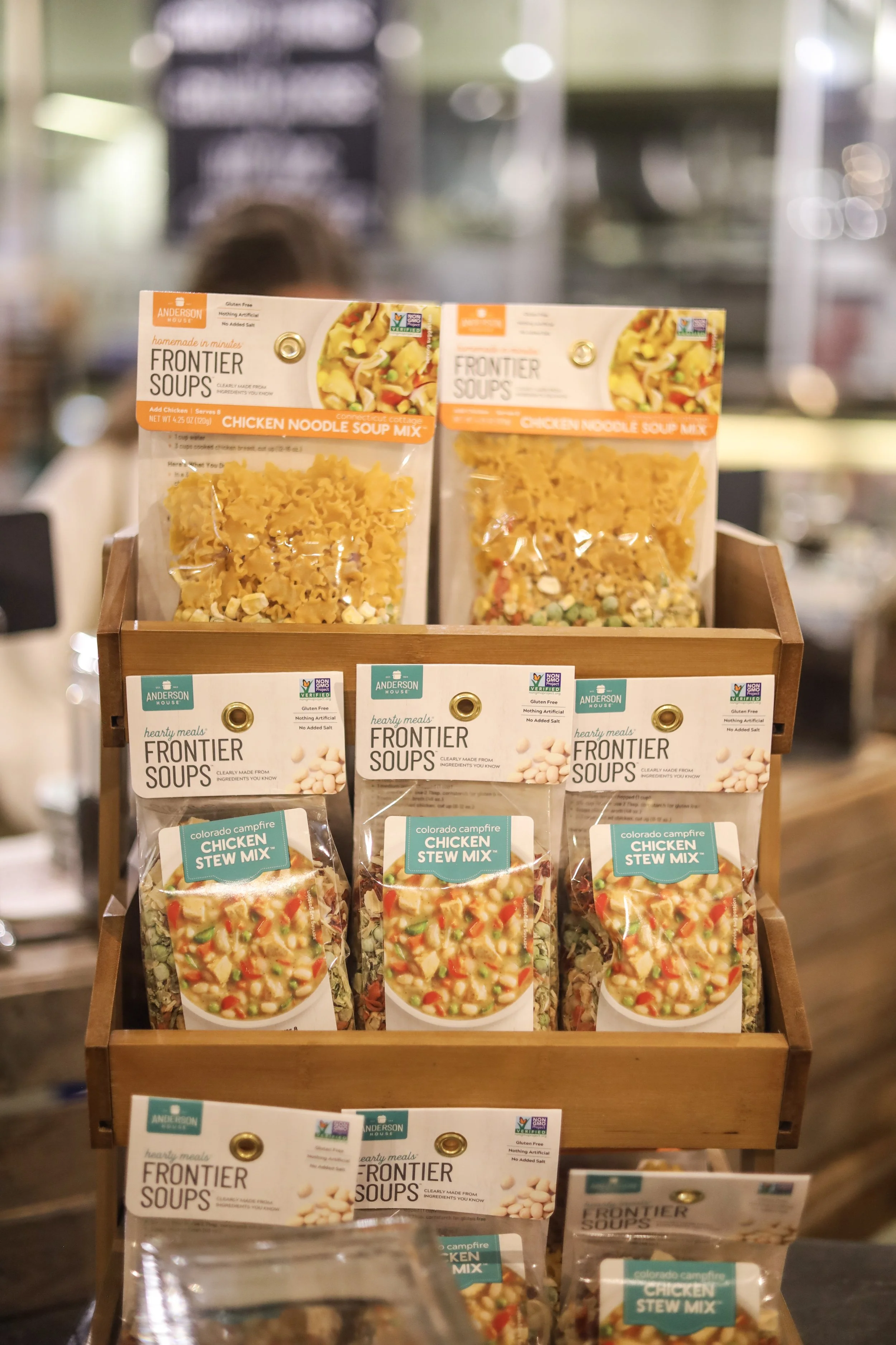 Display of assorted soup mixes on a wooden shelf at a grocery store. The top row has packages of chicken noodle soup mix, and the lower rows have packages of chicken stewed mix. The packaging features colorful images of soup ingredients.