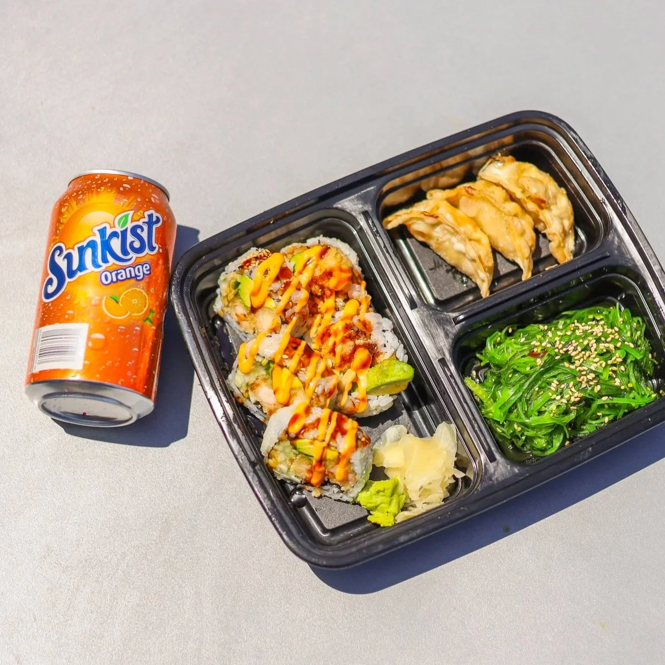 Bento box filled with sushi, potstickers, and seaweed salad on a metal table in the sunshine with a can of soda.