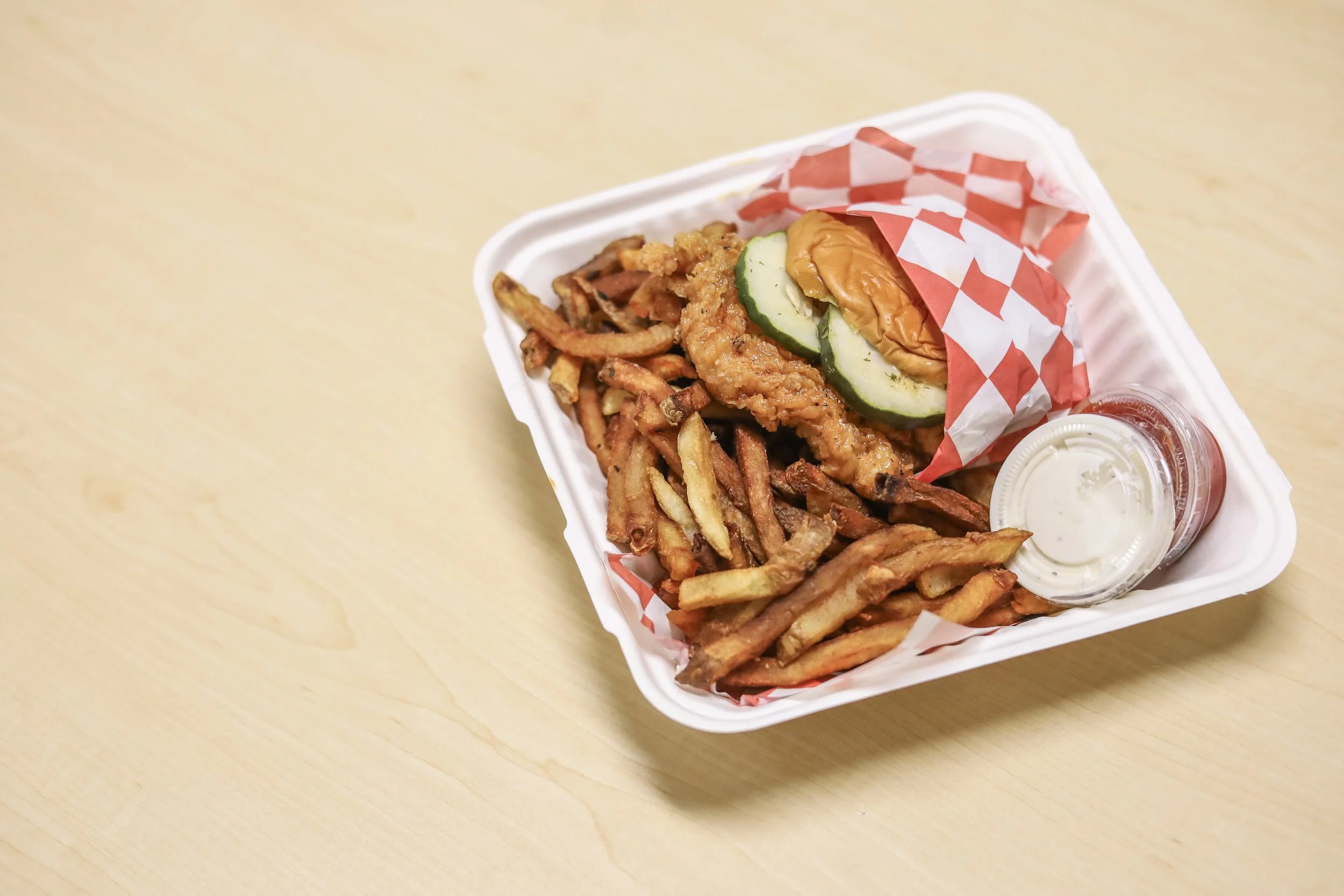 Takeout tray with fried chicken sandwich, seasoned French fries, ketchup, and dipping sauce on a wooden surface.