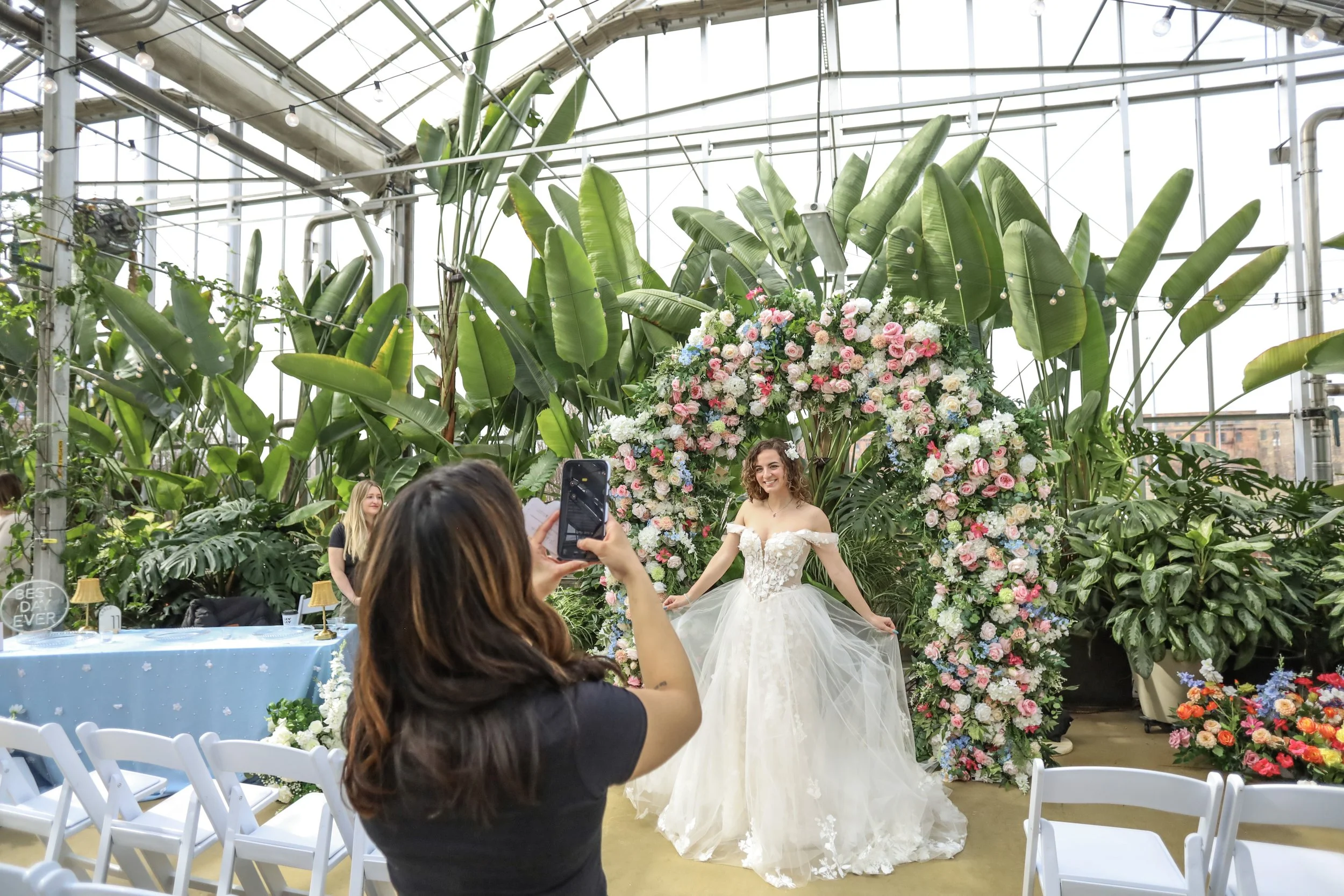 Woman in wedding dress taking a picture with a smartphone in front of a floral arch in a greenhouse.