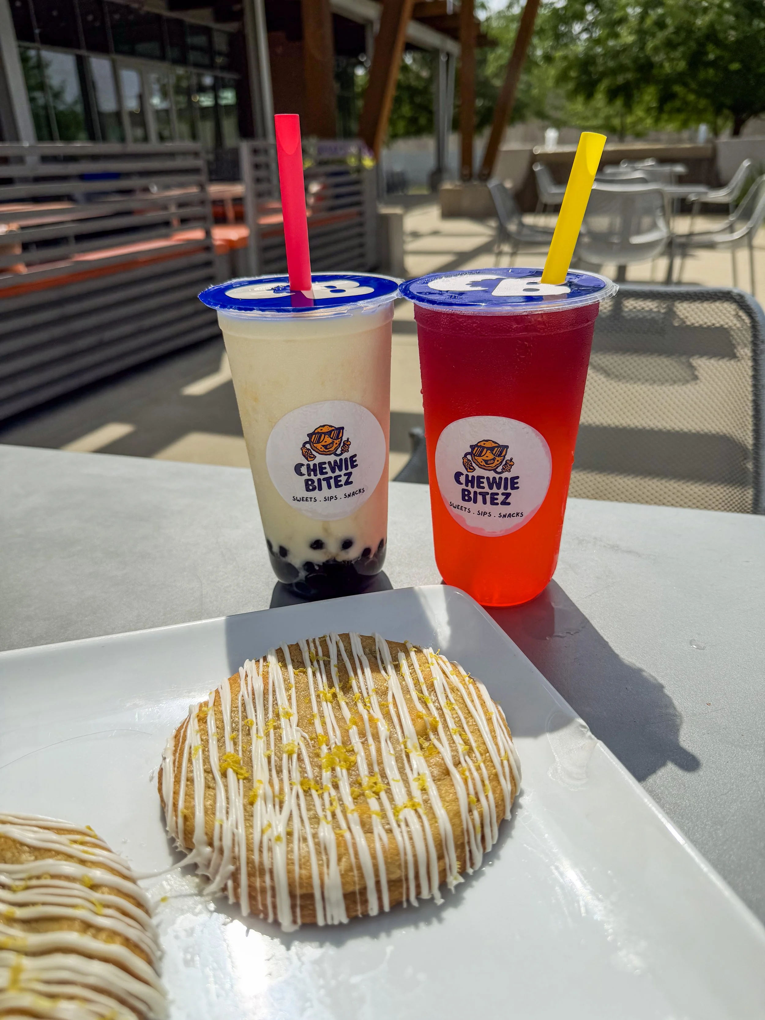 A large stuffed cookie sitting on a plate in the sunshine with two bubble teas in the background