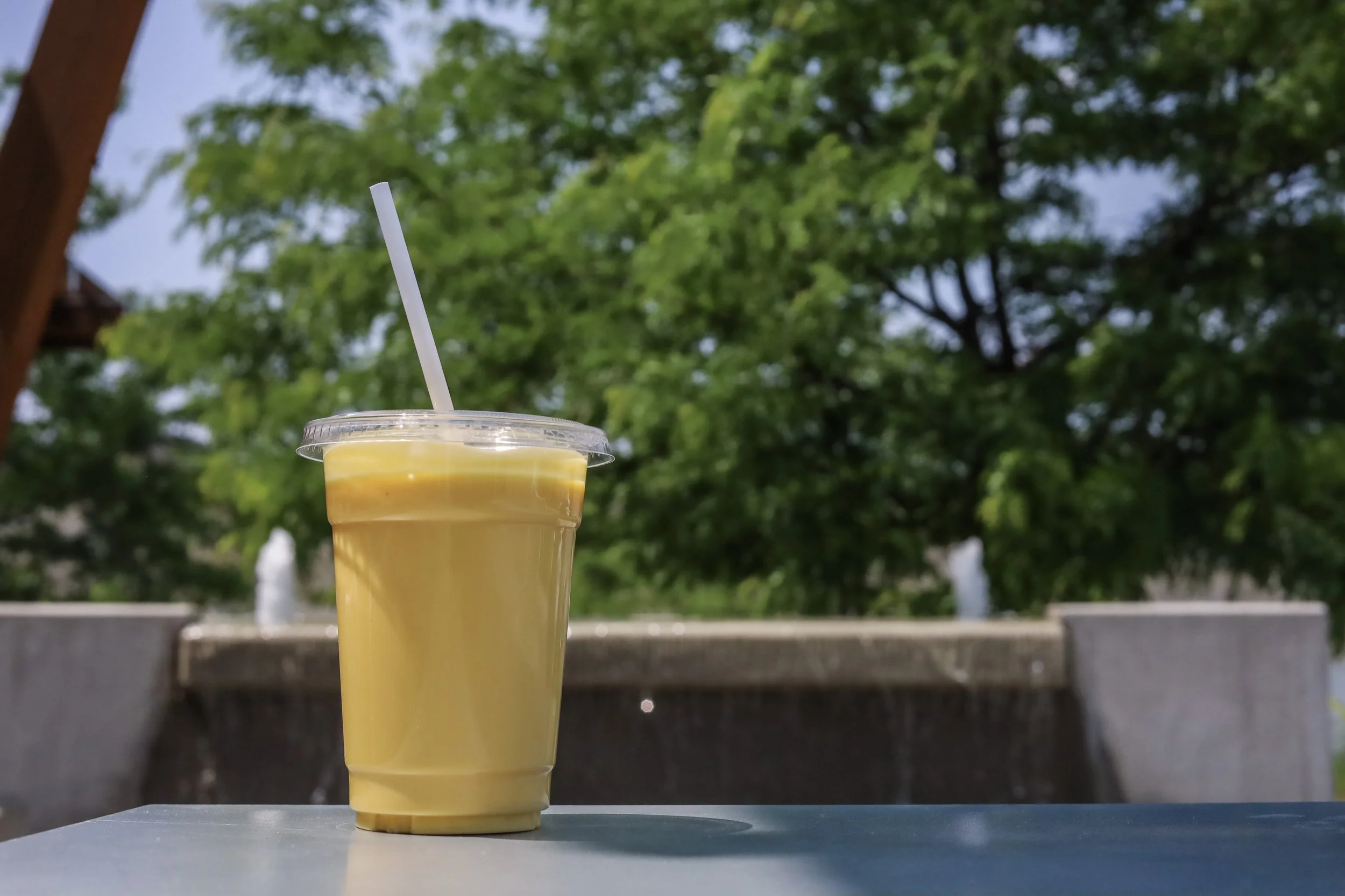 A yellow smoothie in a clear plastic cup with a lid and straw, placed on a table outdoors with green trees and a water fountain in the background.
