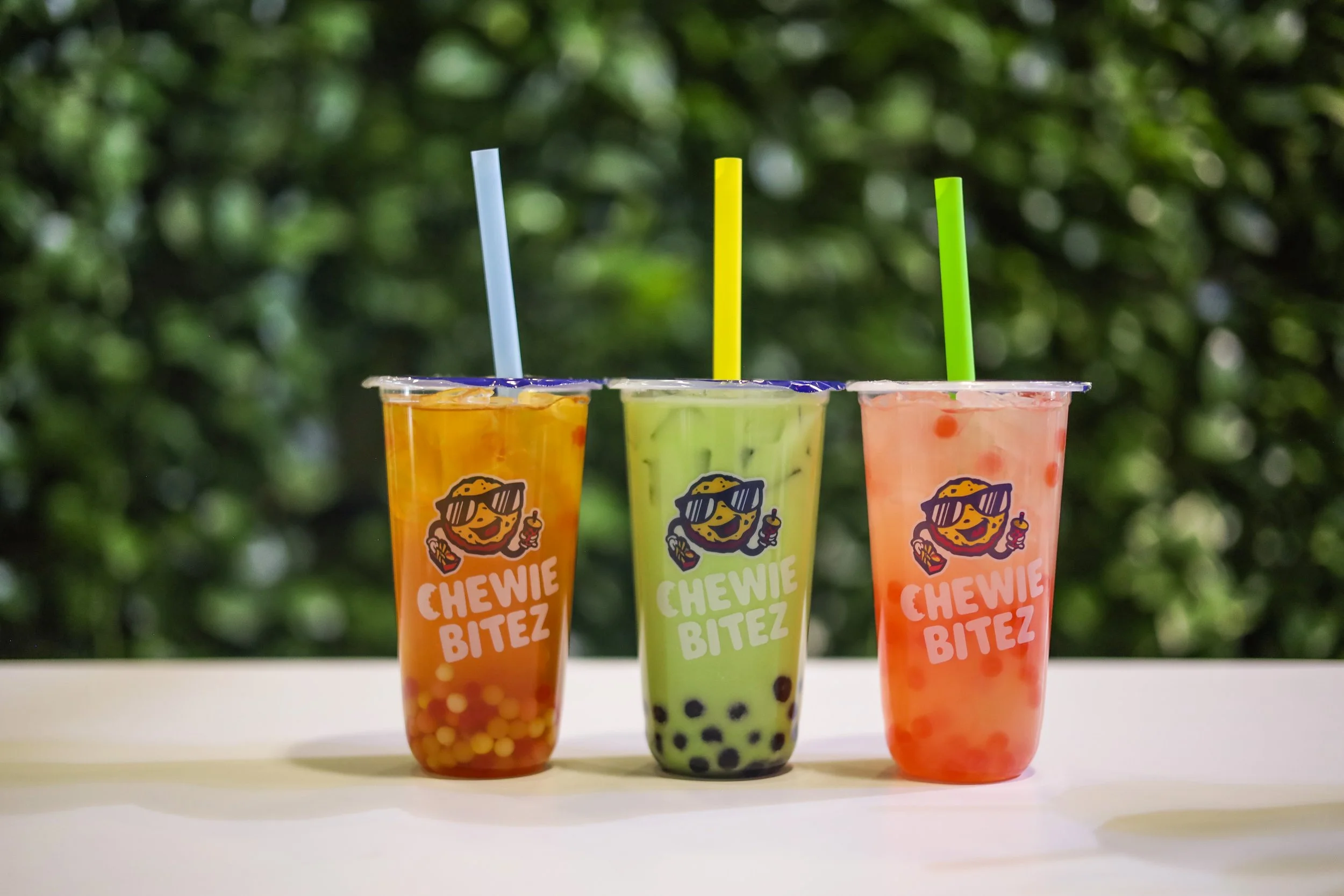 Three colorful bubble tea drinks in clear cups with Chewie Bitez logo, with blue, yellow, and green straws, set on a white surface with a green, leafy background.