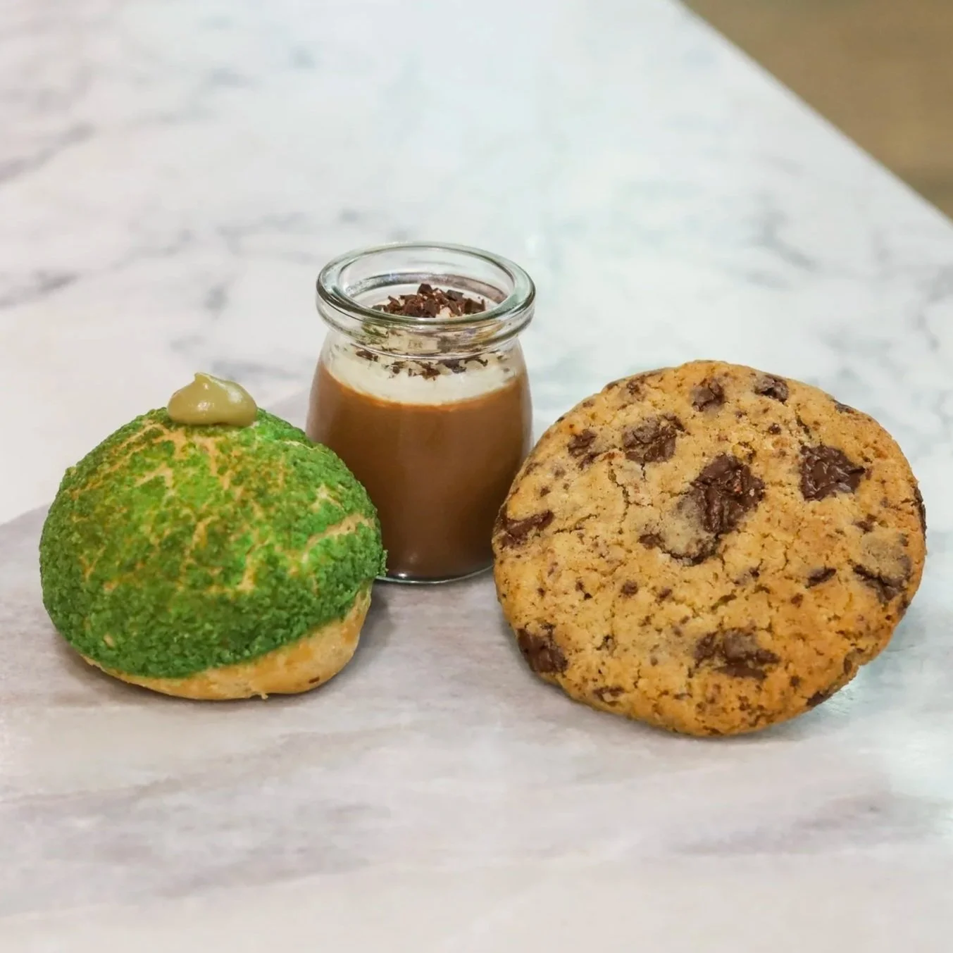 Pistachio cream puff, chocolate mousse cup, and a chocolate chip cookie, laying on marble.