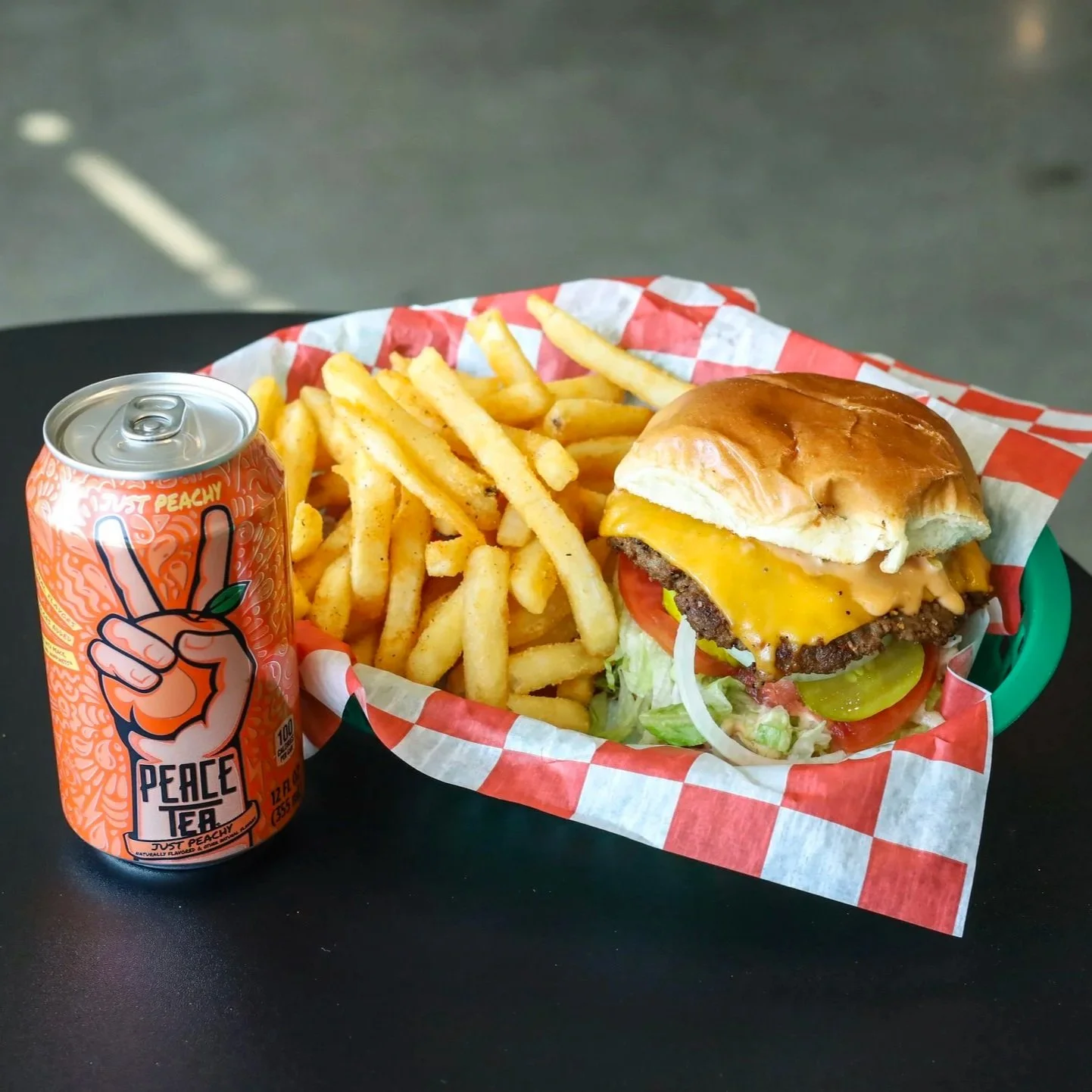 A vegan burger with lettuce, tomato, pickles, and vegan cheese in a bun, served with fries and a can of tea.