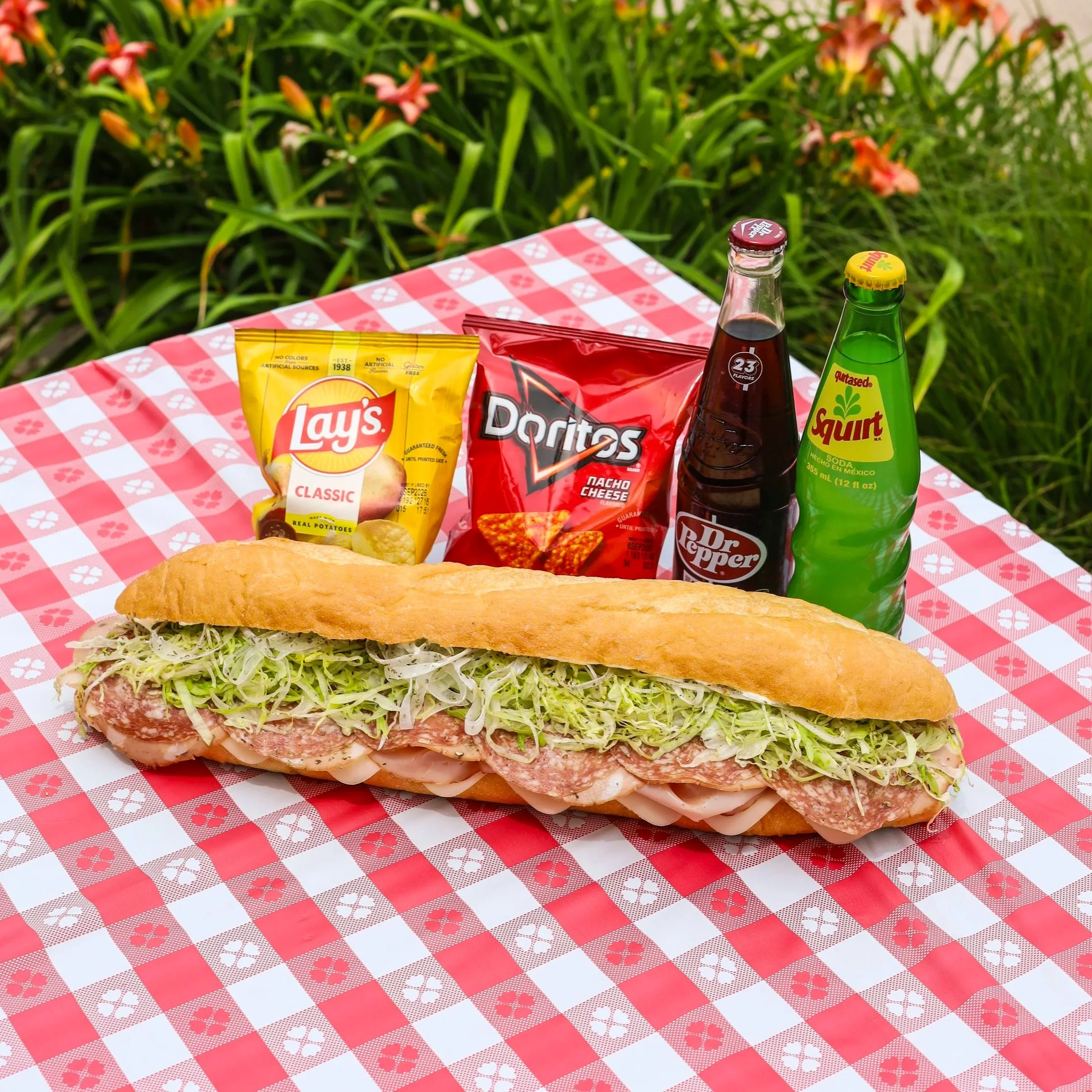 A huge sub sandwich sitting on a tablecloth outside next to two craft sodas and two bags of chips.
