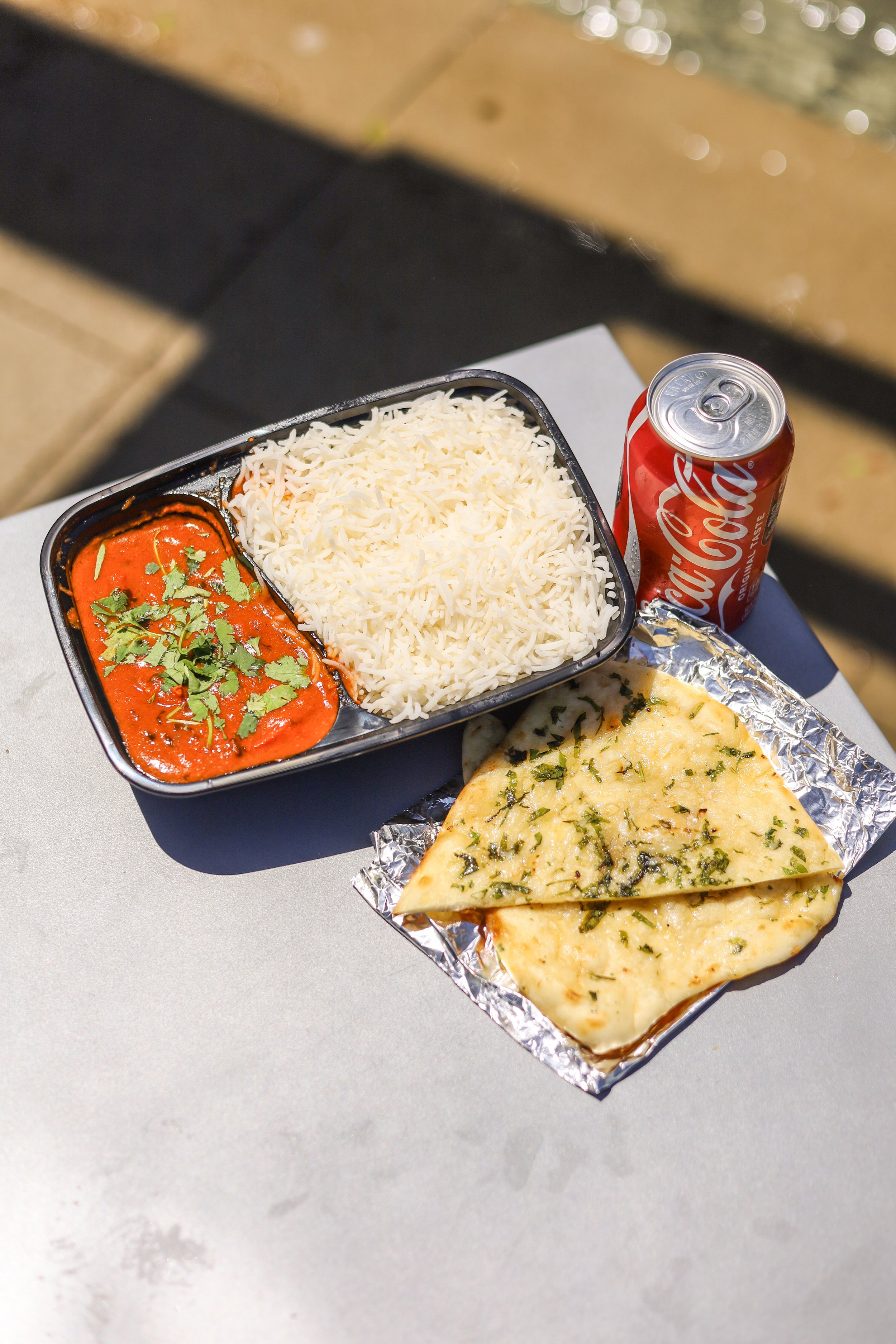 Chicken Tikka Masala and fluffy rice with a side of butter naan and a can of soda on a table in the sunshine.