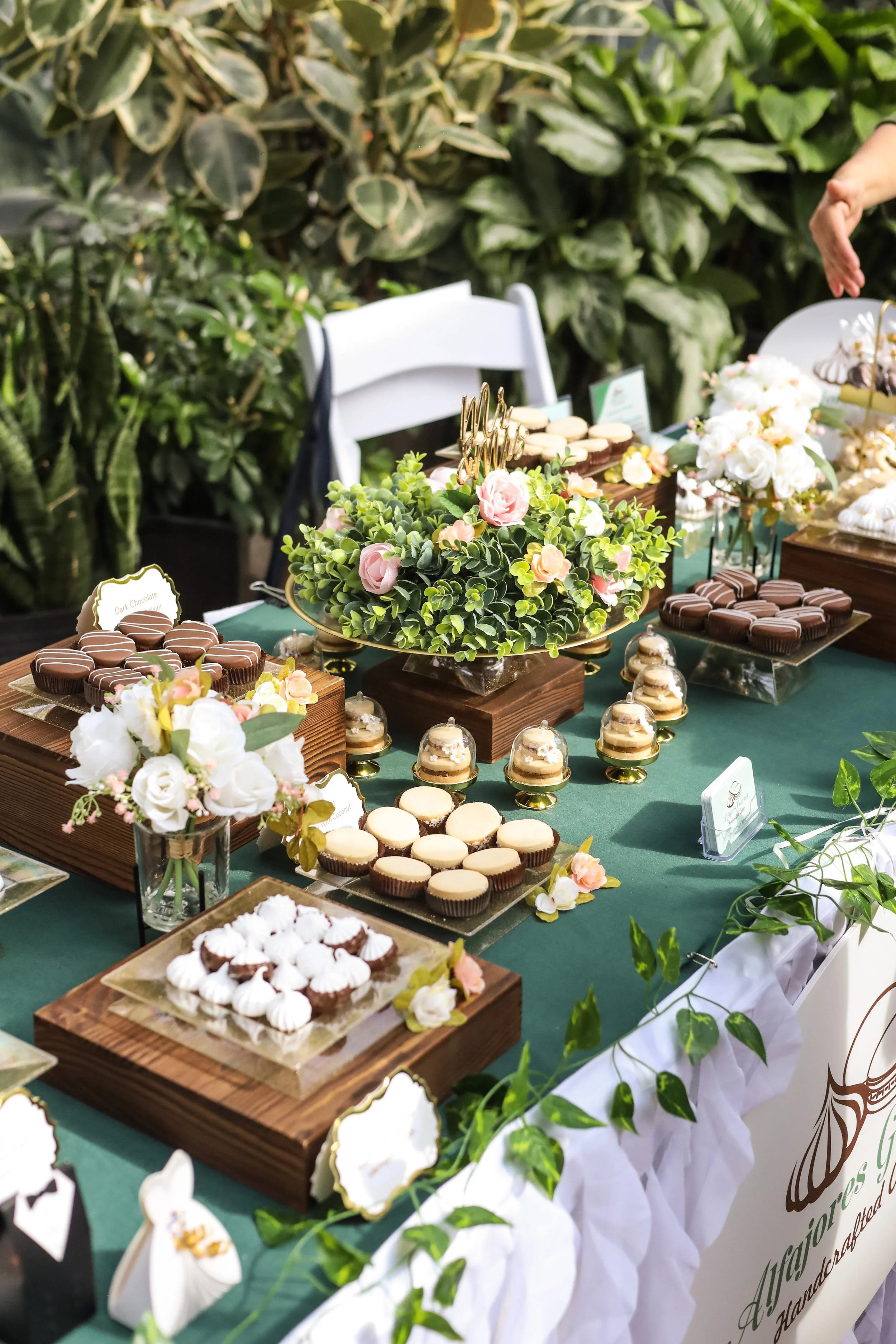 A dessert table with various chocolates, macarons, and pastries, decorated with floral arrangements and greenery at a celebration or event.
