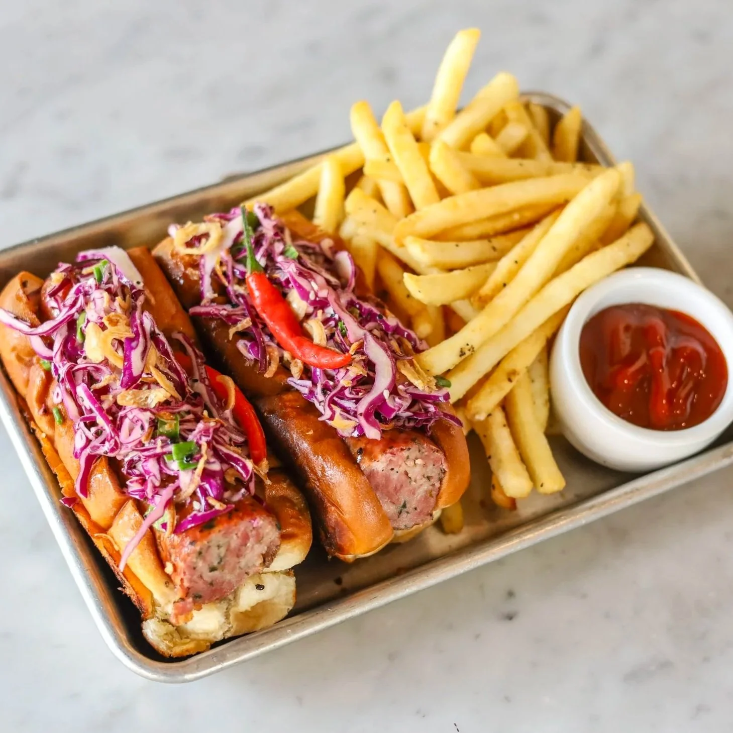 Two grilled Thai sausages in brioche buns topped with slaw and peppers in a tray with fries and spicy ketchup