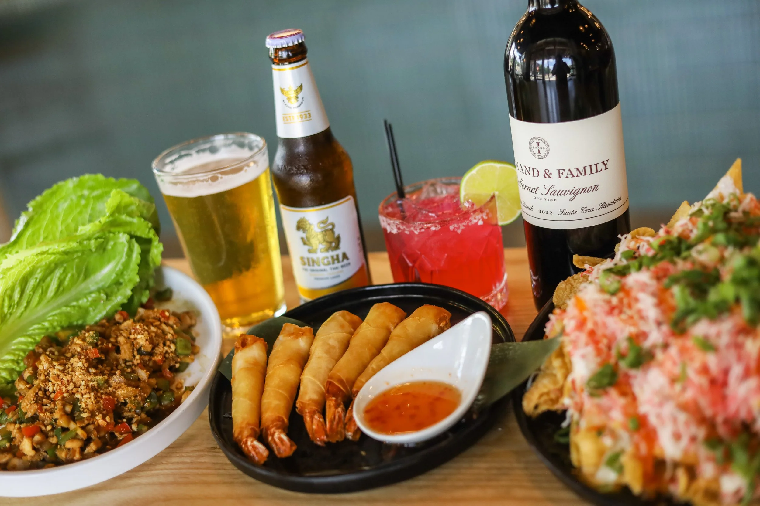 A table filled with colorful Thai food, lettuce wraps, crab cheese nachos, and alcoholic beverages.