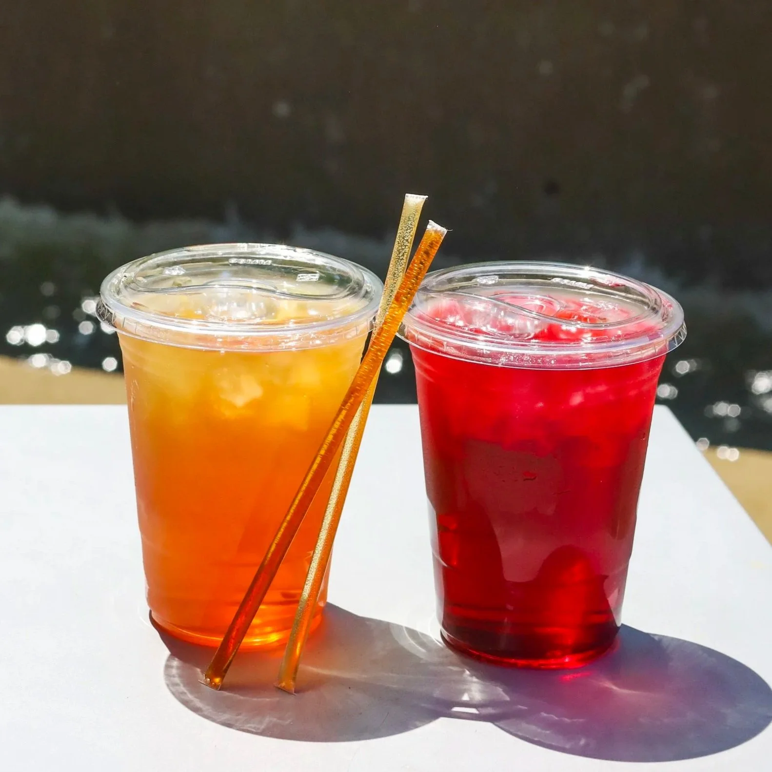 Two iced teas glistening in the sun with honey straws on a table by the water.