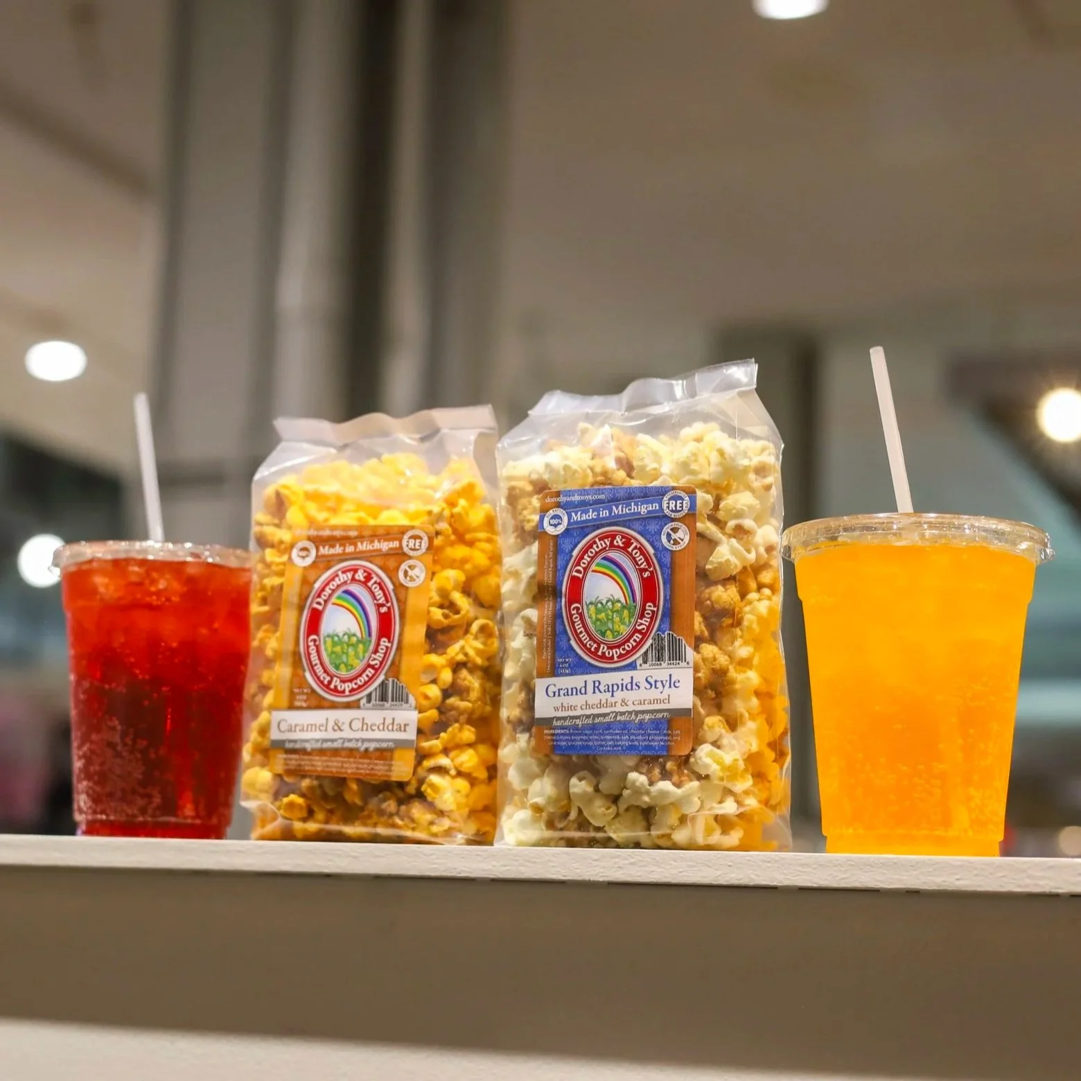 Two bags of popcorn on a ledge with two brightly colored fountain sodas.