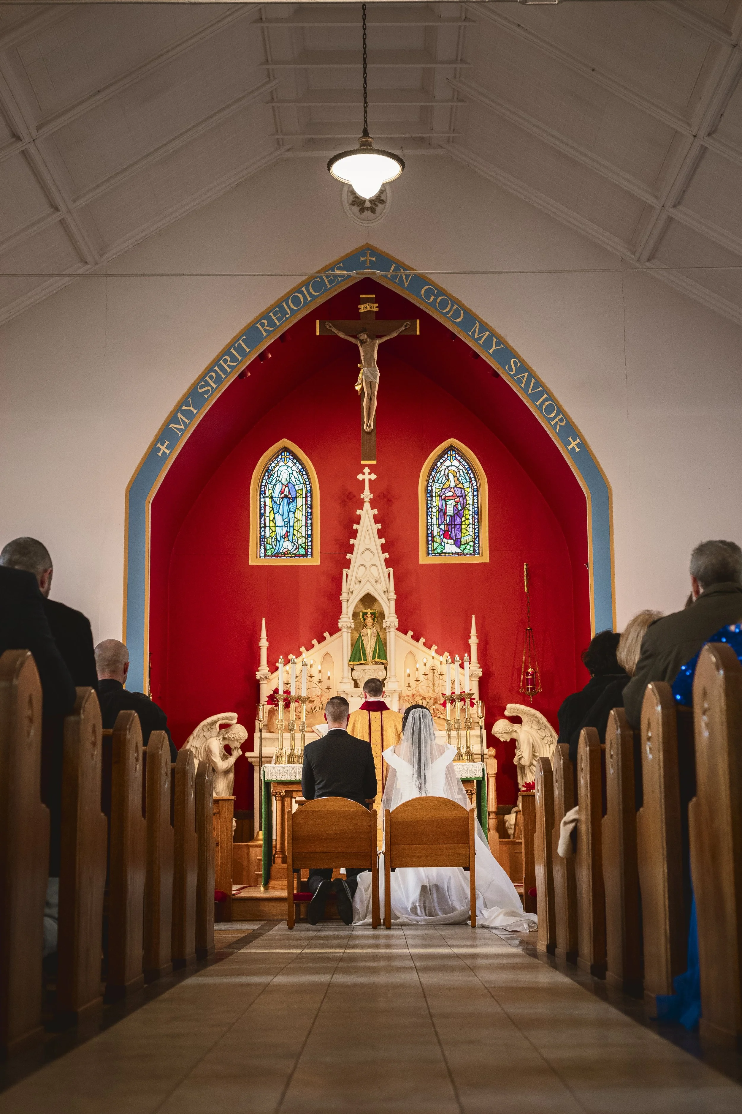 A wedding ceremony taking place inside a church with stained glass windows, a crucifix, and an altar decorated with candles, flowers, and religious statues, with guests seated in pews.