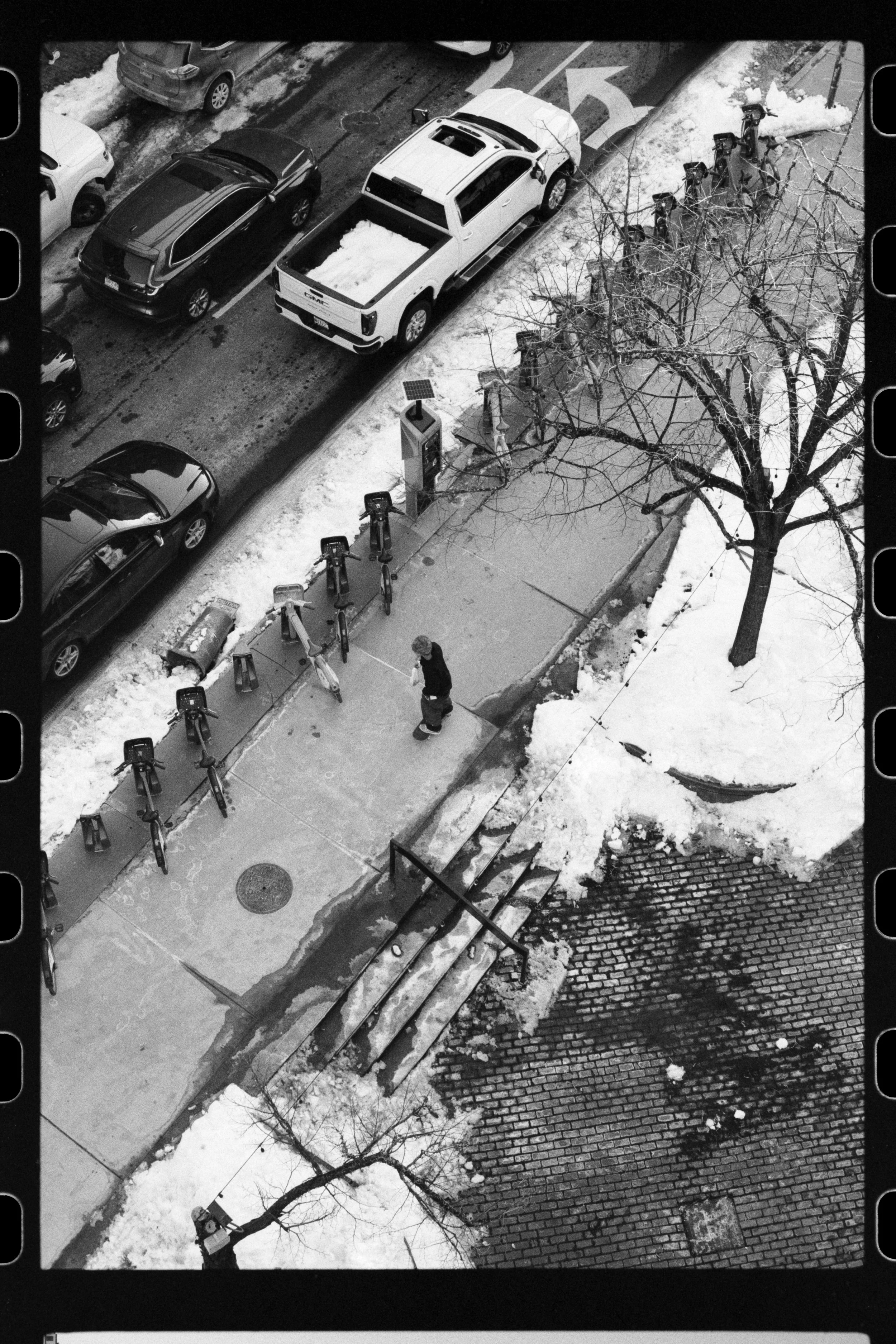Black and white photo of a snow-covered city sidewalk with a row of bike-sharing stations, a leafless tree, a parking lot with several parked cars, and a small person walking on the sidewalk.
