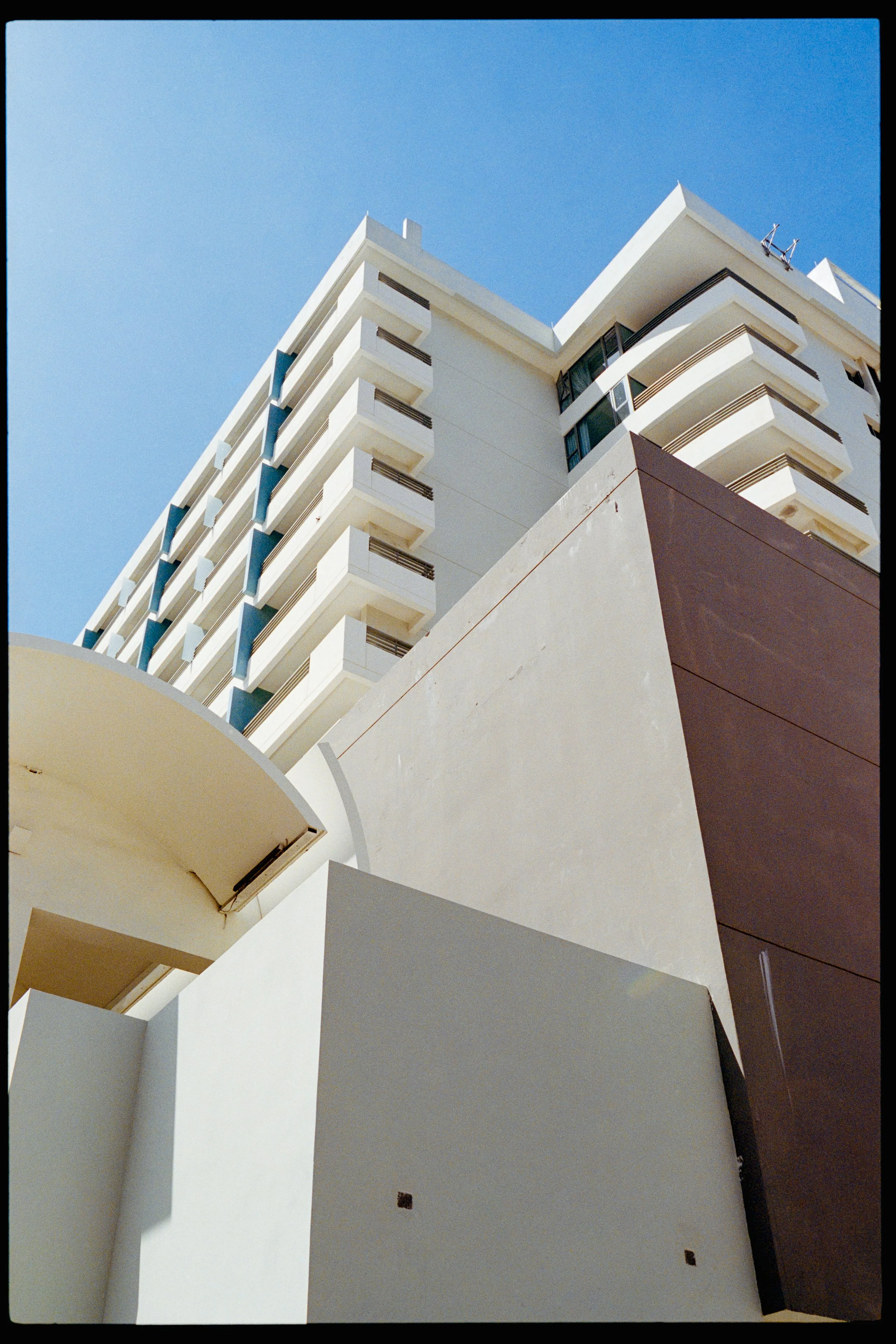 Low-angle view of a modern white building with multiple balconies and glass windows against a clear blue sky.