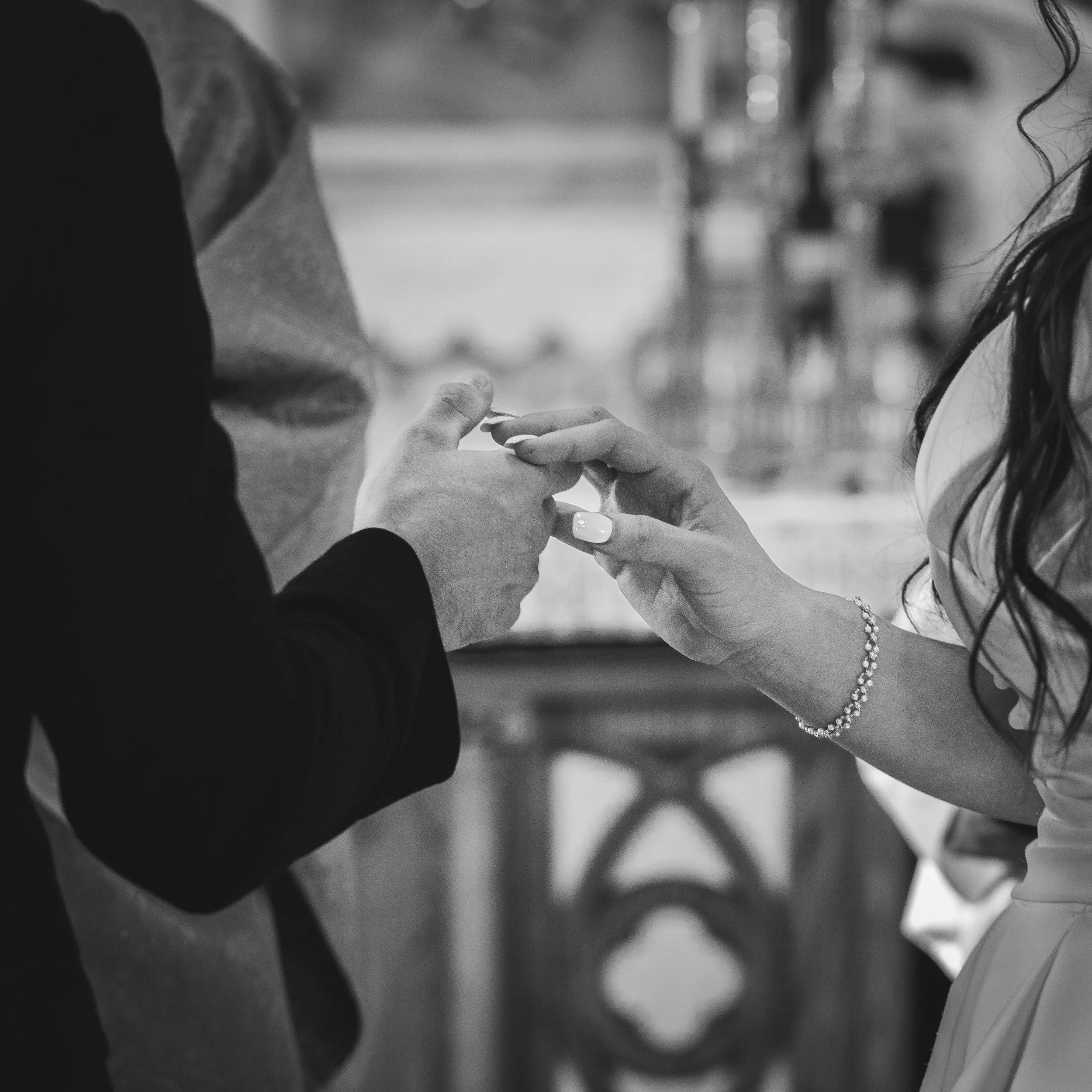 A close-up black and white photo of a couple exchanging rings during a wedding ceremony, with the man wearing a suit and the woman in a dress, holding hands over an altar or podium.