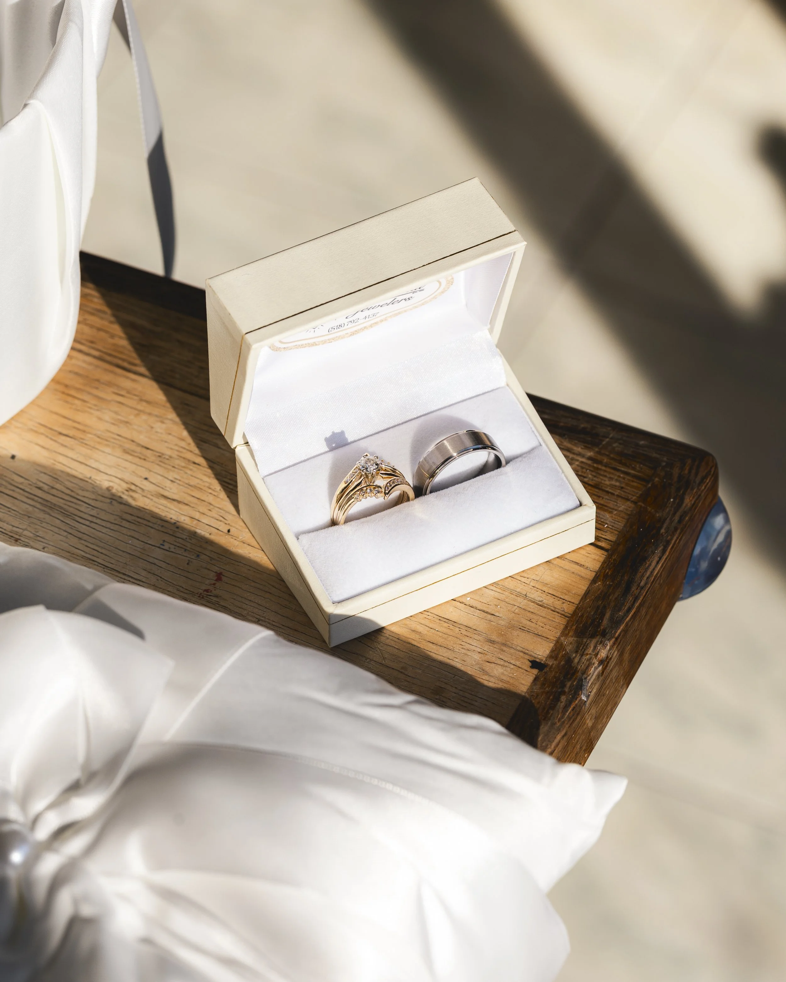 A jewelry box with a gold engagement ring and a silver wedding band inside, placed on a wooden table with sunlight shining on it.