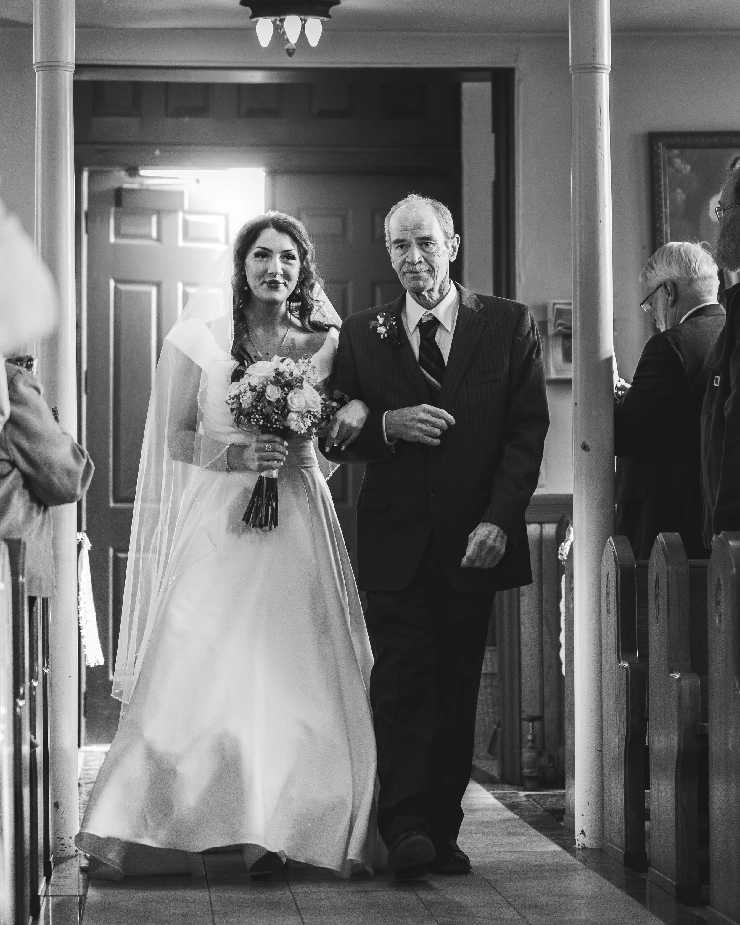 A bride in a wedding dress holding a bouquet, being escorted down the aisle by an older man at a wedding ceremony.