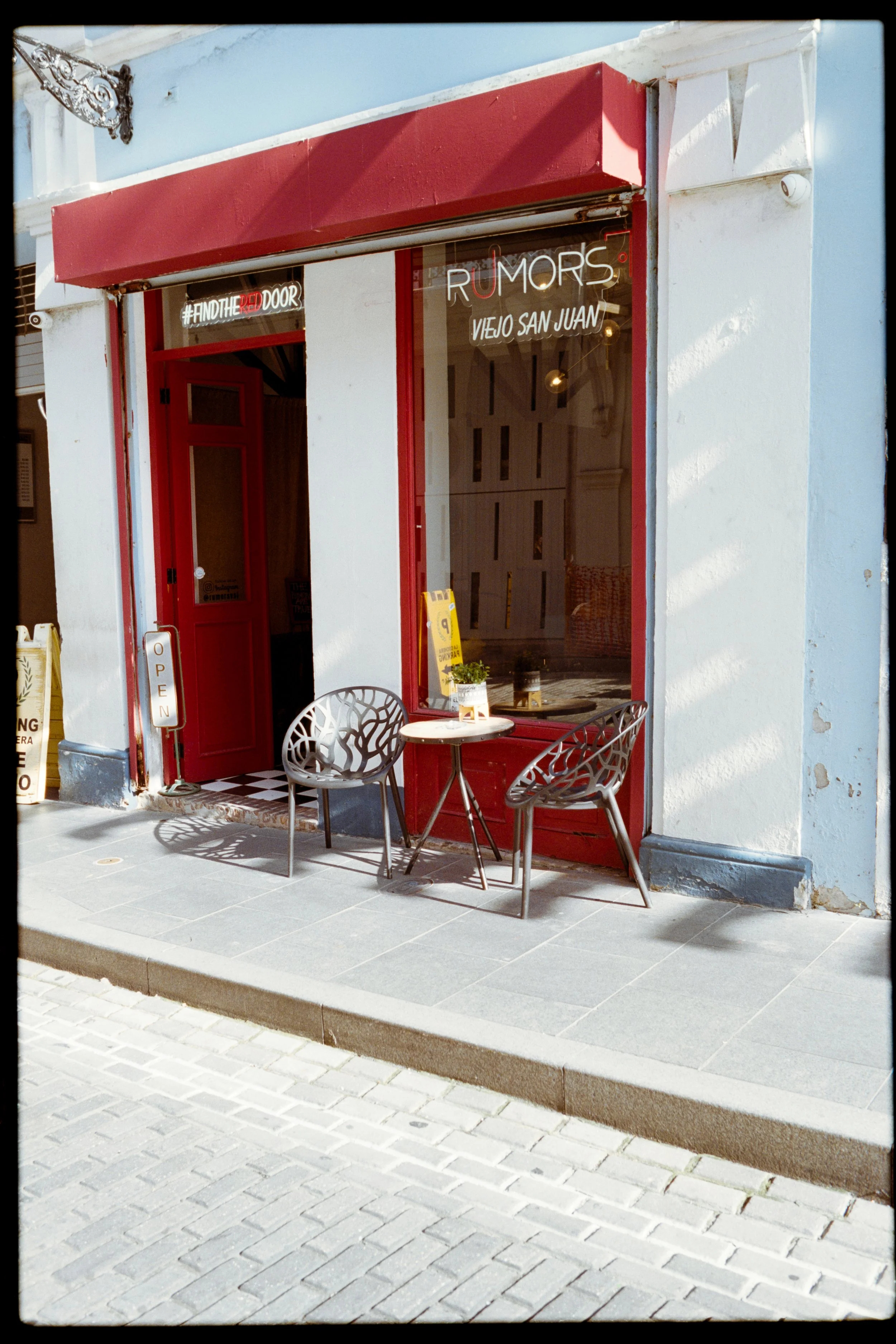 A storefront with a red awning and white walls, featuring a neon sign that reads '#FINDTHE' with unreadable text, a large window with signs that say 'RUMORS' and 'VIEJO SAN JUAN', and two metal chairs with a small table outside on a tiled sidewalk.