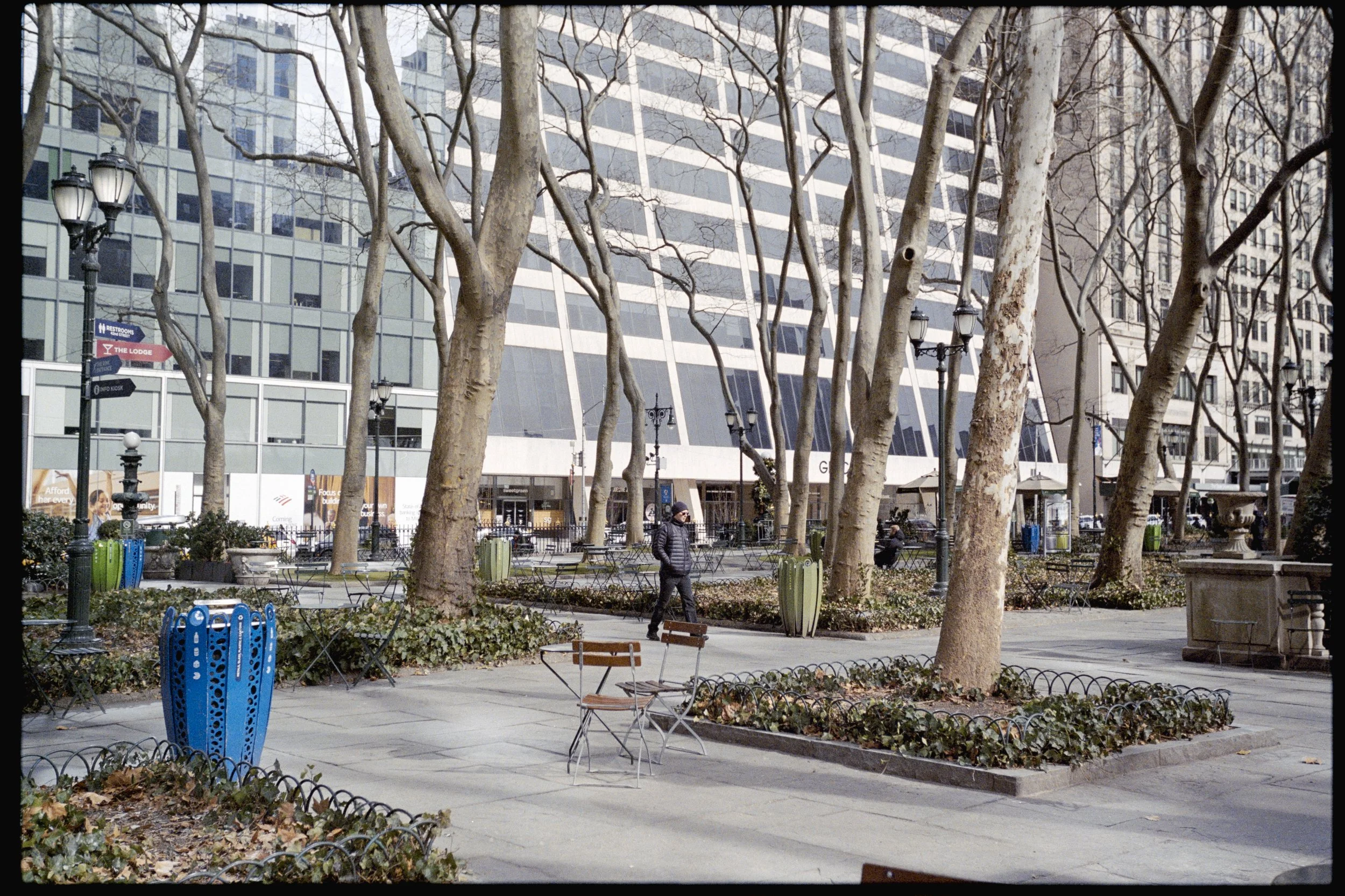 A city park with leafless trees, benches, colorful trash bins, and vintage-style street lamps in front of a modern glass building with a grid pattern. A person is walking.
